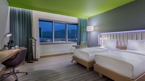 A hotel room with two beds, a desk with a chair, a lamp, and a view of the mountains through the window. - Park Inn by Radisson Samsun (Tekkekoy)