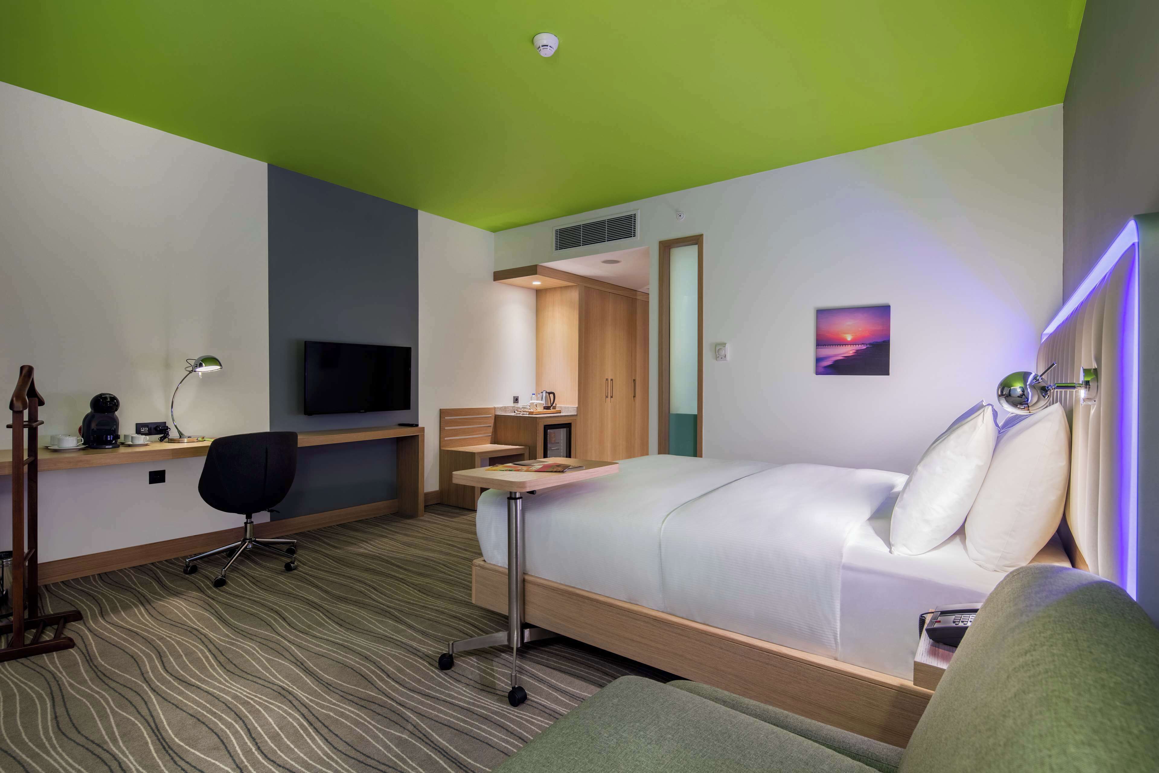 A hotel room with a bed, desk, chair, TV, and a painting on the wall.