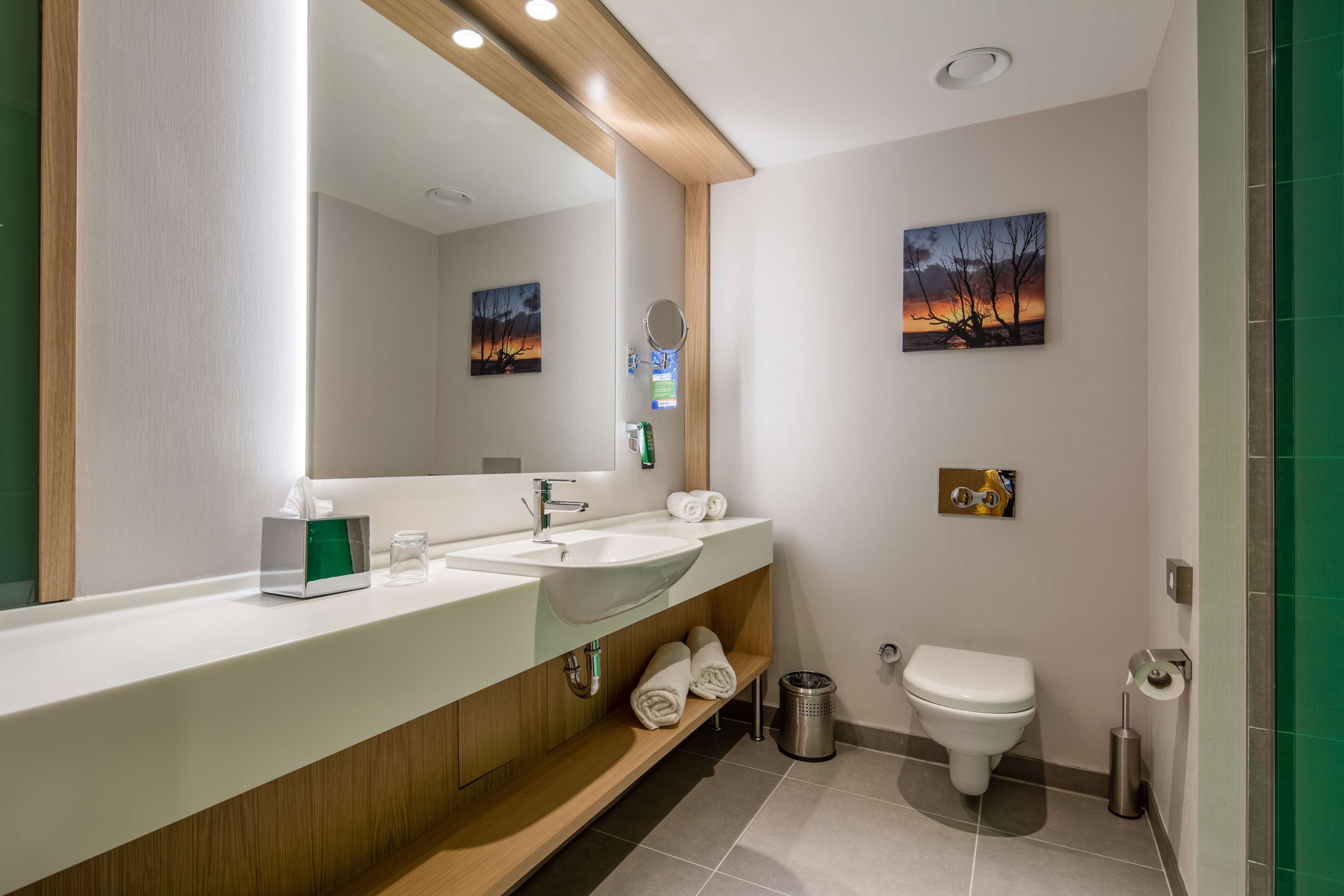 A modern bathroom with a large mirror, two sinks, a toilet, and a trash can.
