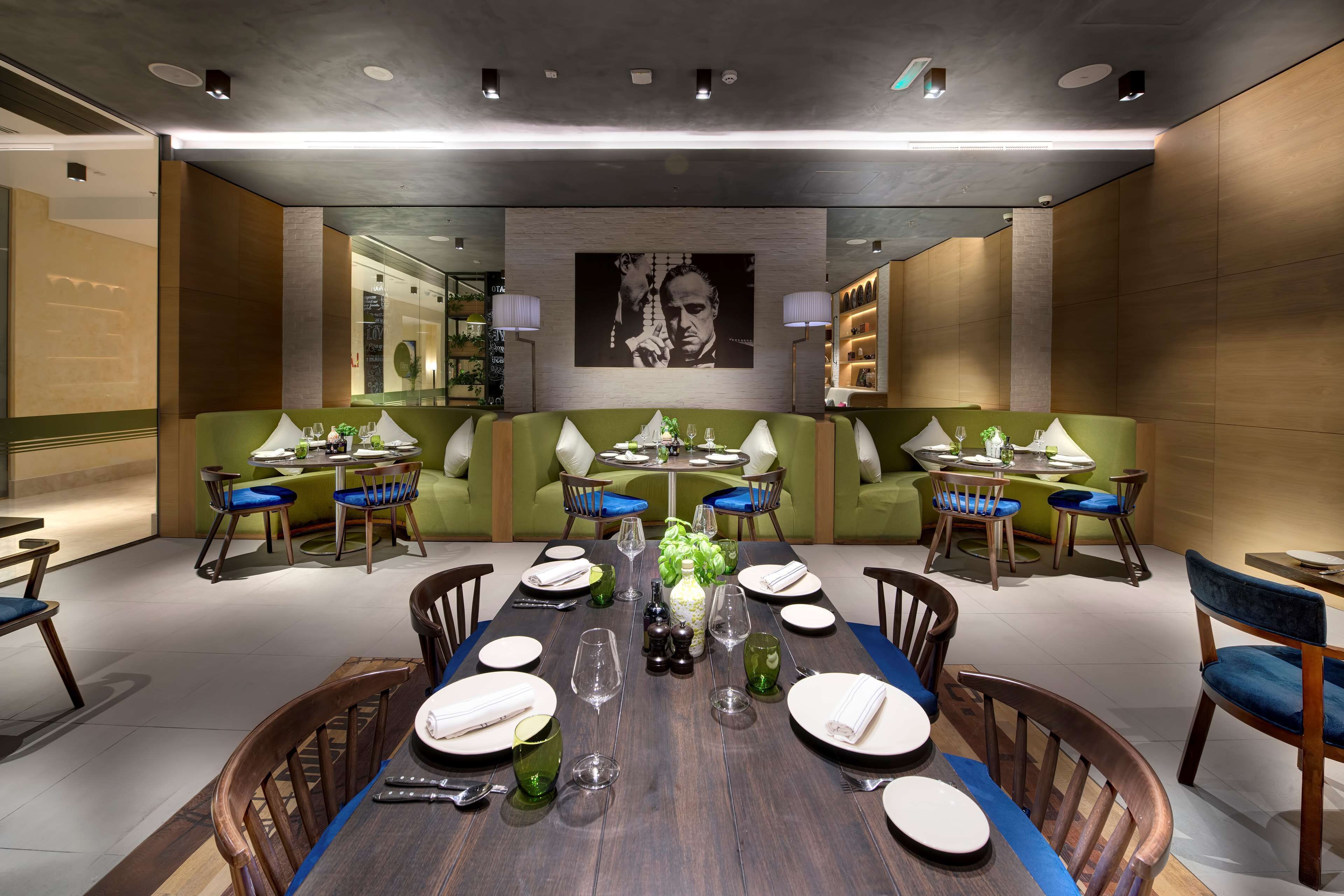 Een eetruimte met een lange tafel voor zes personen, omringd door groene, beklede zithoeken en een grote muurschildering.