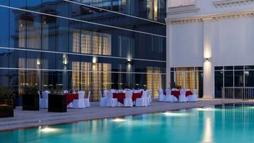 Un área de comedor junto a la piscina con mesas dispuestas para una comida, rodeada por un edificio moderno con amplias ventanas y una piscina.