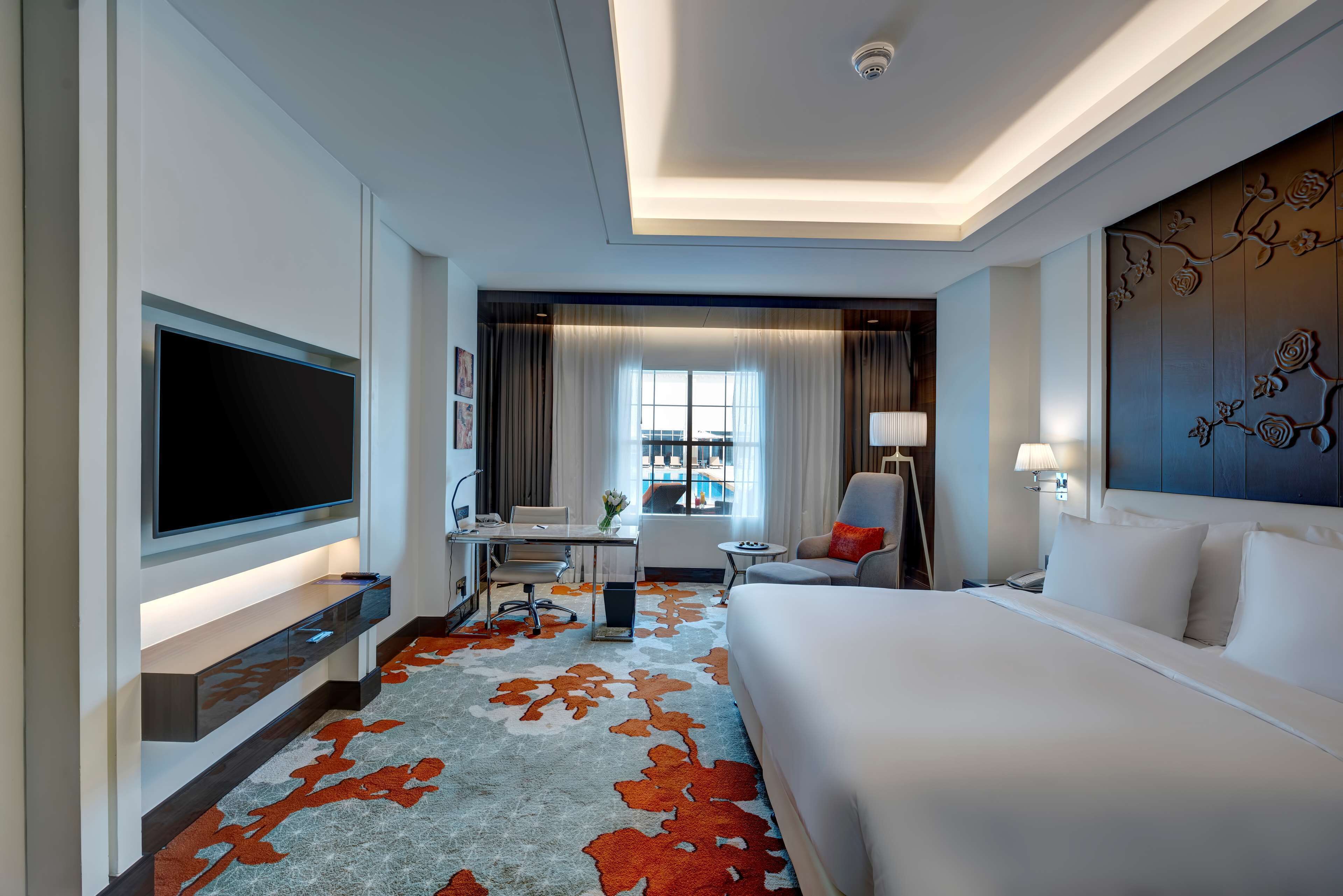 Hotelkamer met een groot bed, een flatscreen televisie, een bureau met stoel, een klein tafeltje en een raam met gordijnen.