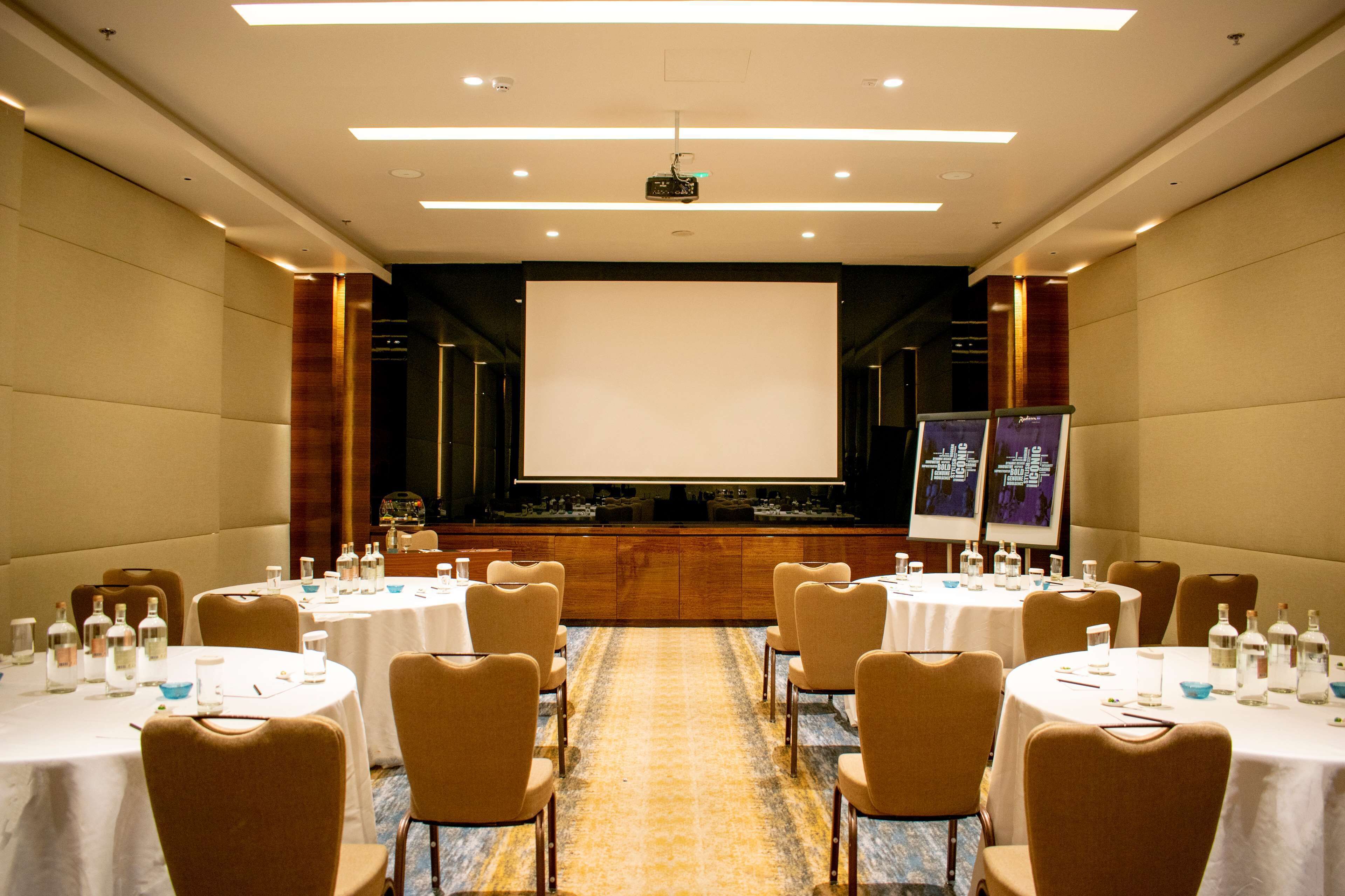 Un salón de conferencias con mesas redondas, sillas, una pantalla de proyector y una pantalla de presentación.