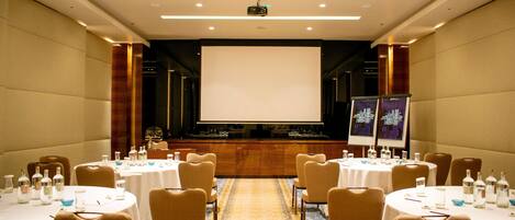 Un salón de conferencias con mesas redondas, sillas, una pantalla de proyector y una pantalla de presentación.