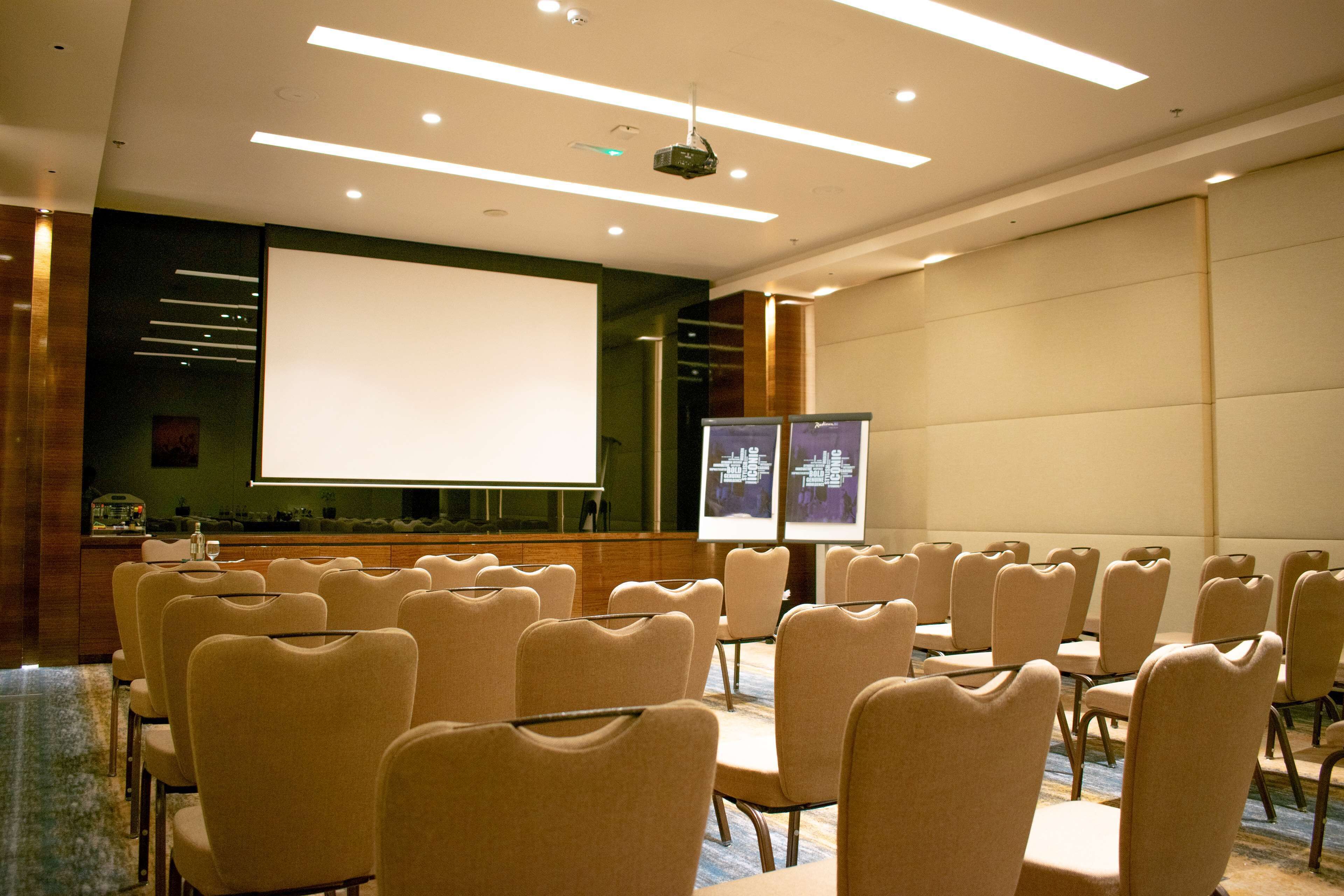 A conference room with rows of beige chairs, a large white projection screen, and two smaller screens on stands.