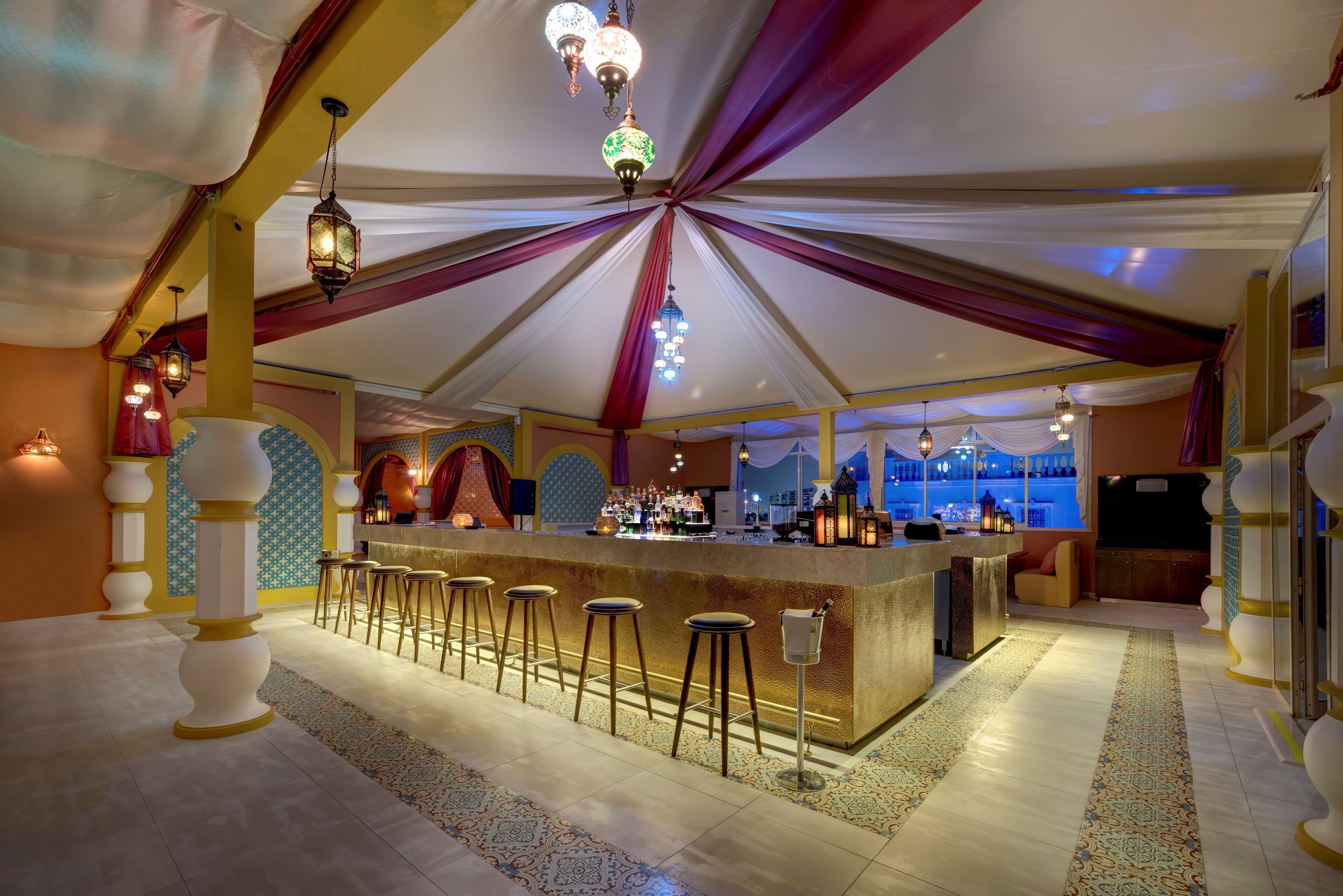 A bar area with a long counter, multiple stools, and hanging lights.