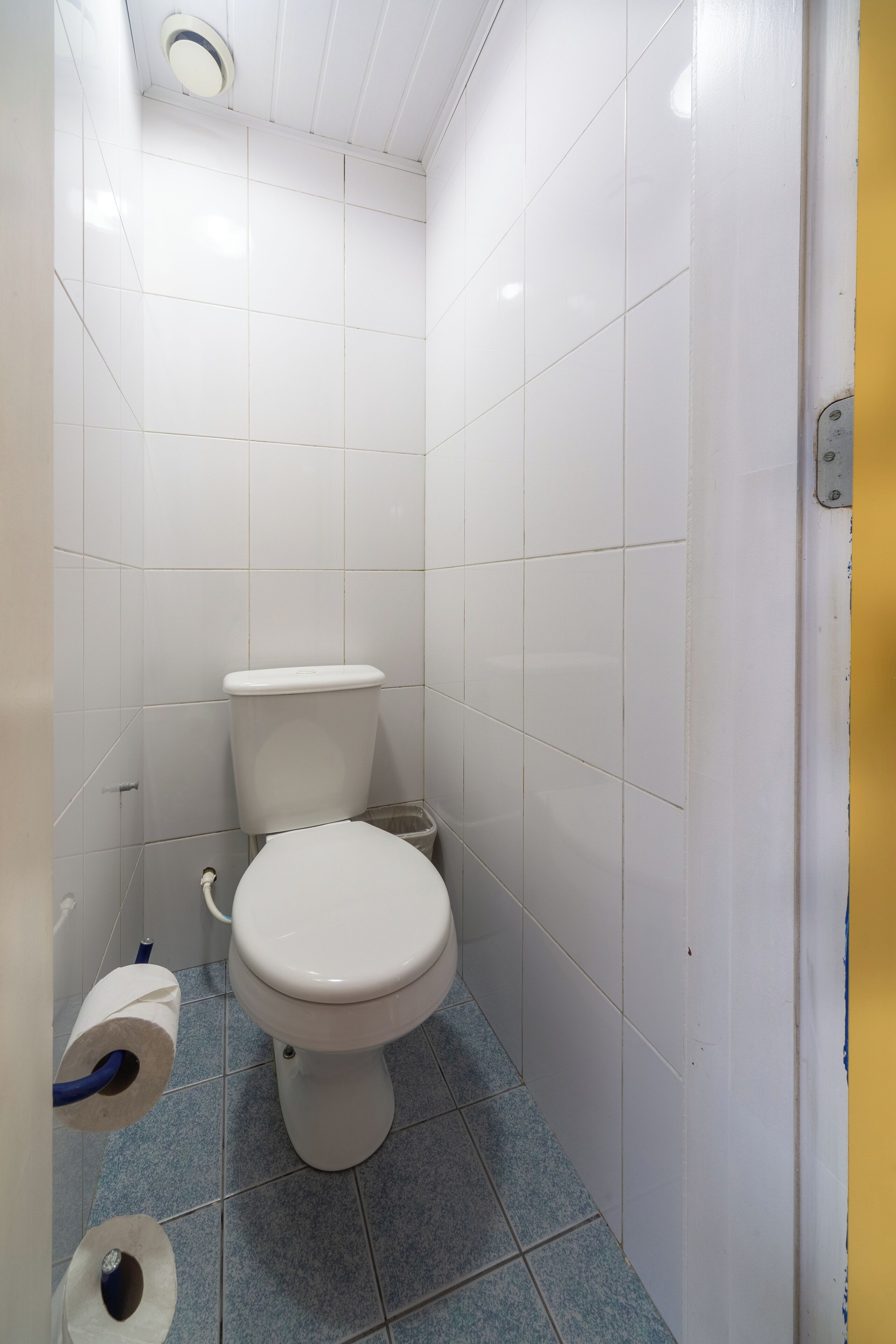 Un baño pequeño y limpio, con azulejos blancos, un inodoro y un rollo de papel higiénico en el suelo.