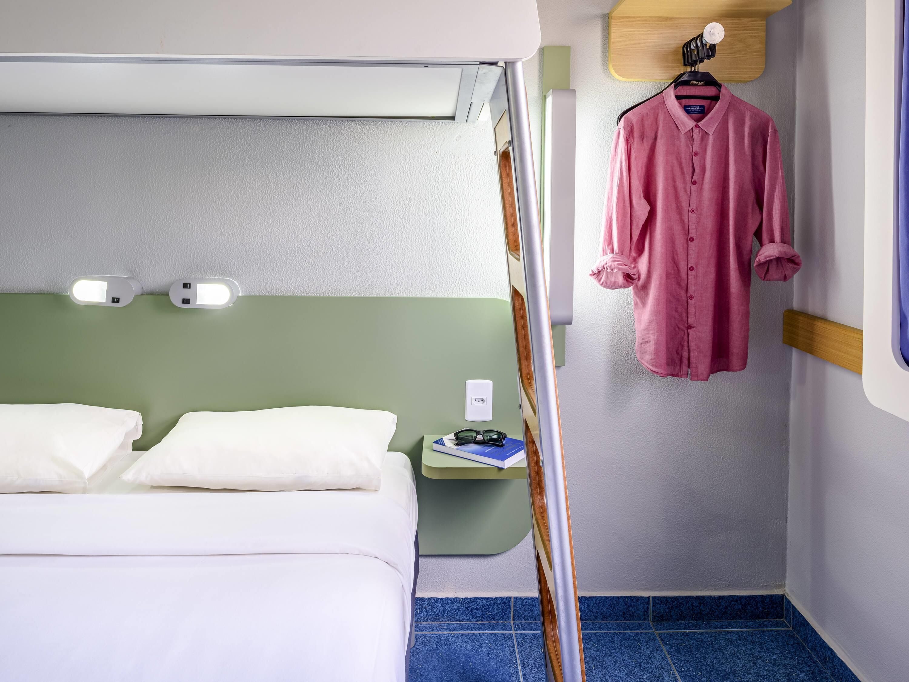 A bedroom with a bed, pillows, a bedside table with glasses, a hanging garment, and a wall-mounted light fixture.