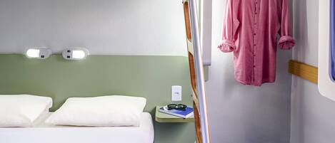 A bedroom with a bed, pillows, a bedside table with glasses, a hanging garment, and a wall-mounted light fixture.
