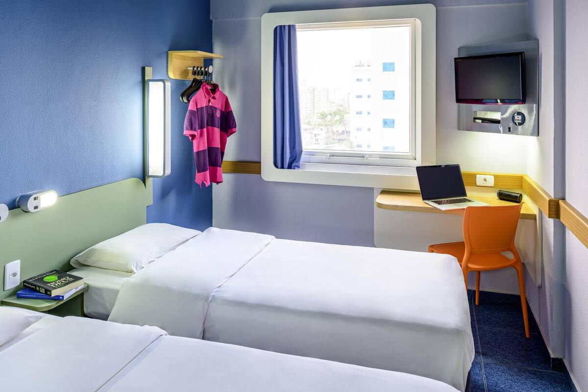 A hotel room with two beds, a desk with a laptop, a chair, a TV, and a window with a view of buildings.