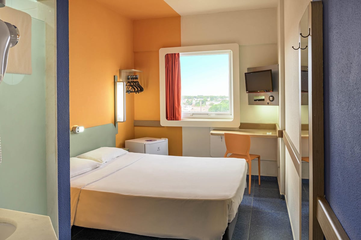 A hotel room with a bed, a desk with a chair, a television, a window with curtains, and a small heater.