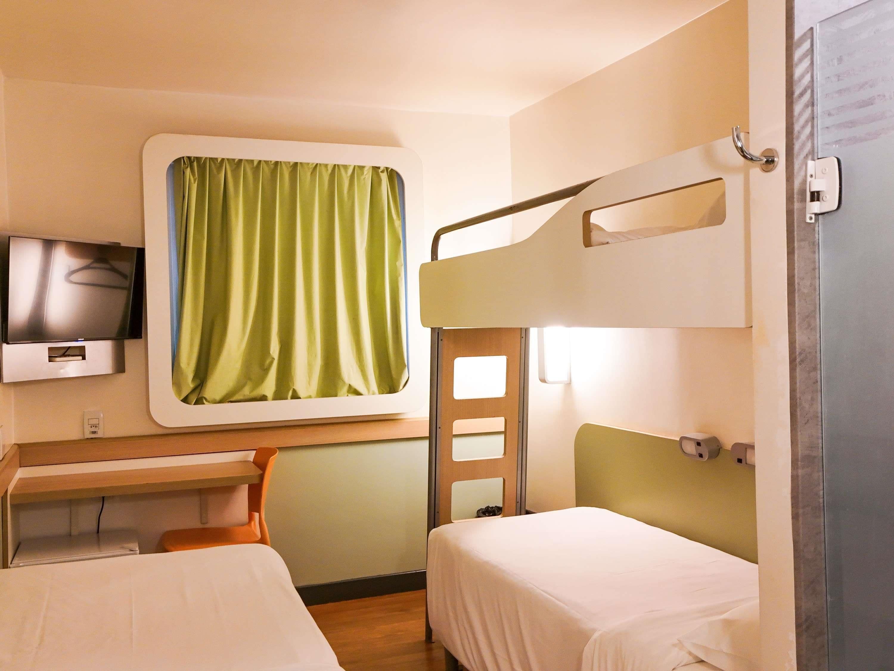 A hotel room with a bunk bed, a desk with a chair, a television, and a window with curtains.