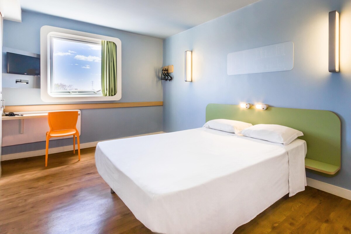 A hotel room with a bed, a desk, and a chair. There is a window with curtains, a wall-mounted light, and a flat-screen TV.