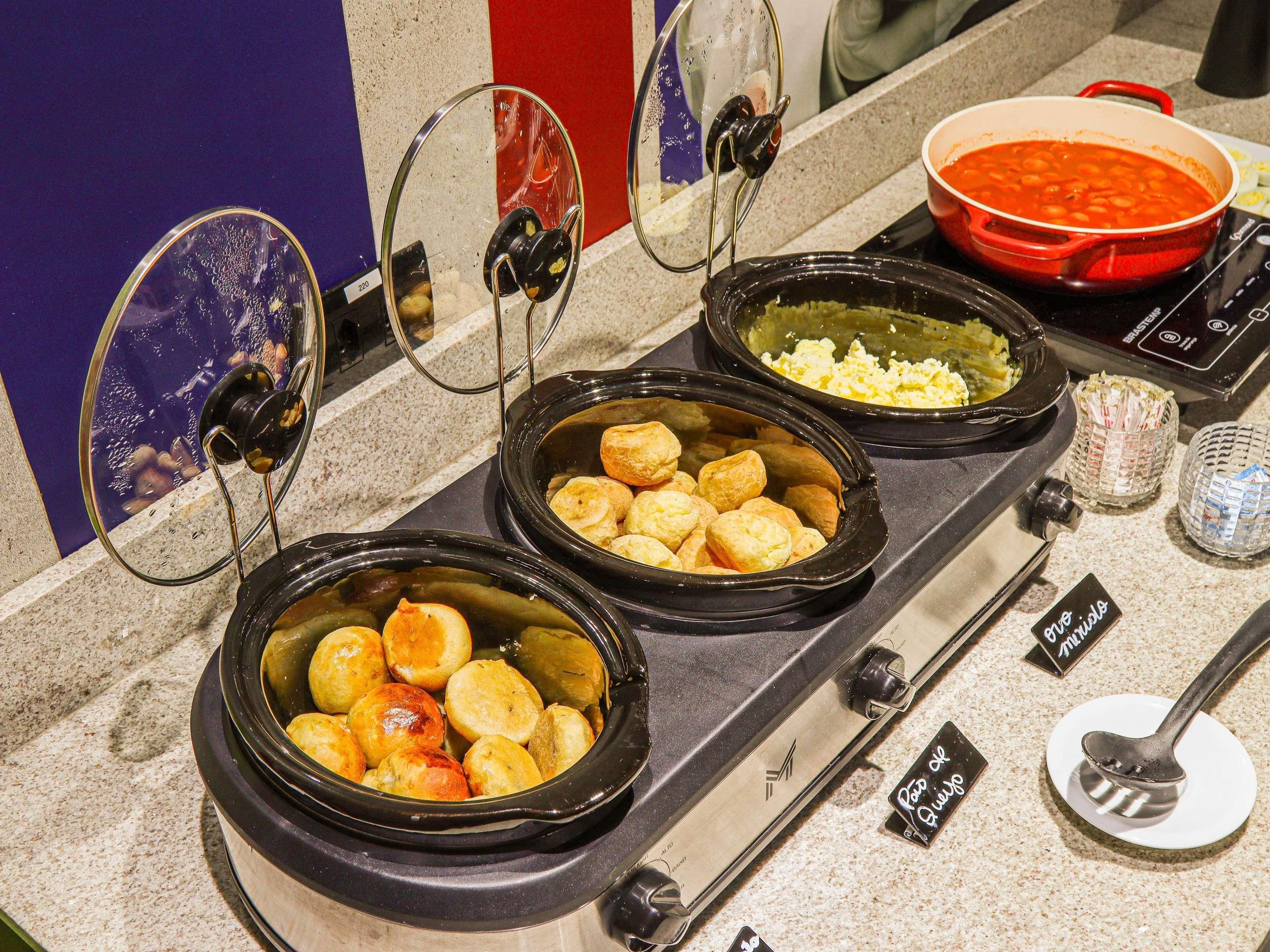 Een keuken met meerdere slowcookers gevuld met verschillende gerechten, een rode pan op het fornuis, en een glas water.