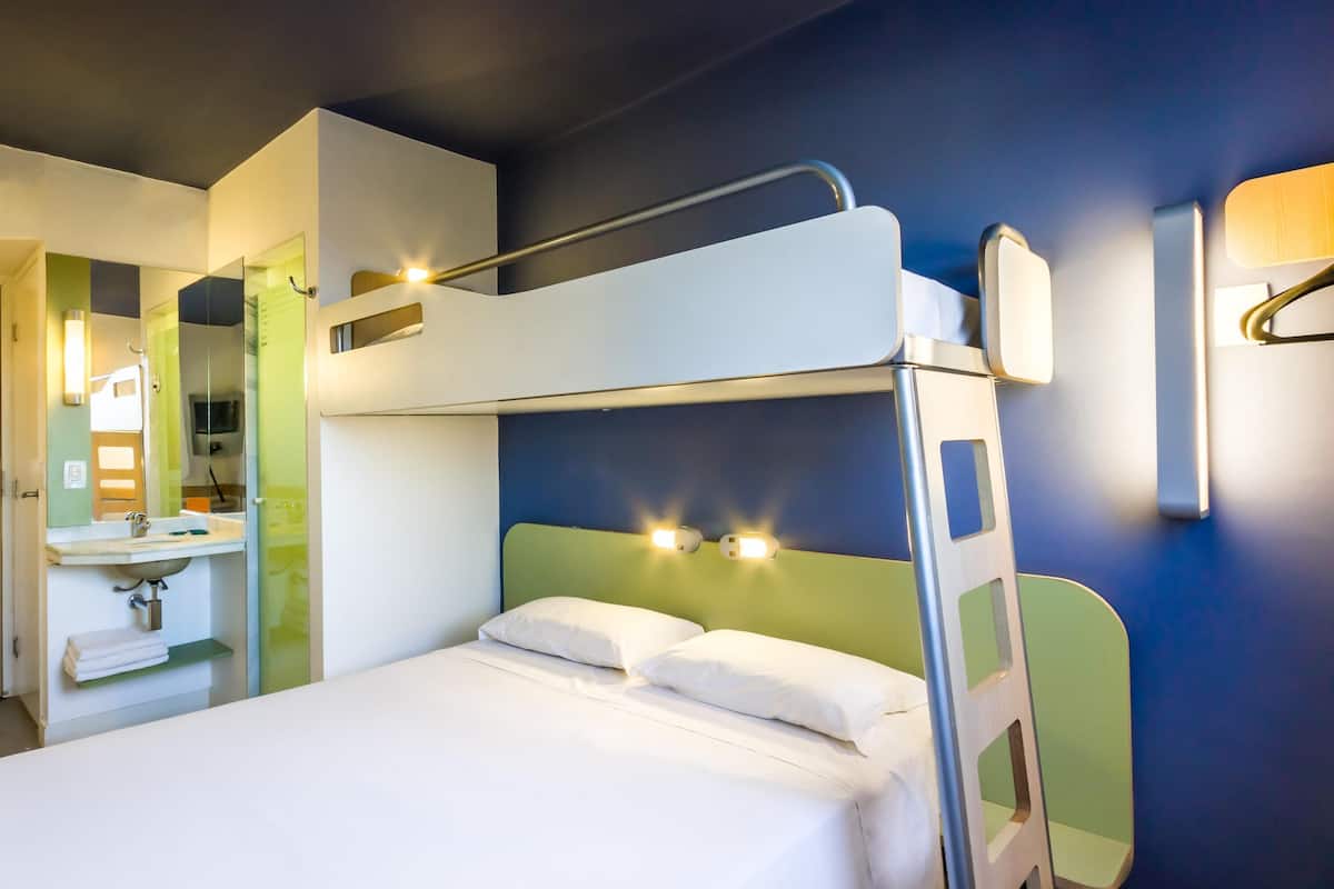 A bunk bed with a white mattress and pillows, a bedside table, a mirror, and a sink with a towel rack.