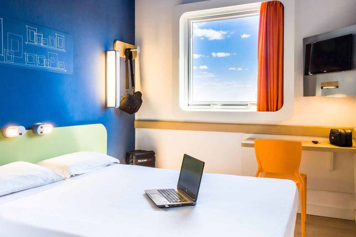 Habitación de hotel con cama, escritorio, computadora portátil, televisor y ventana con cortinas.