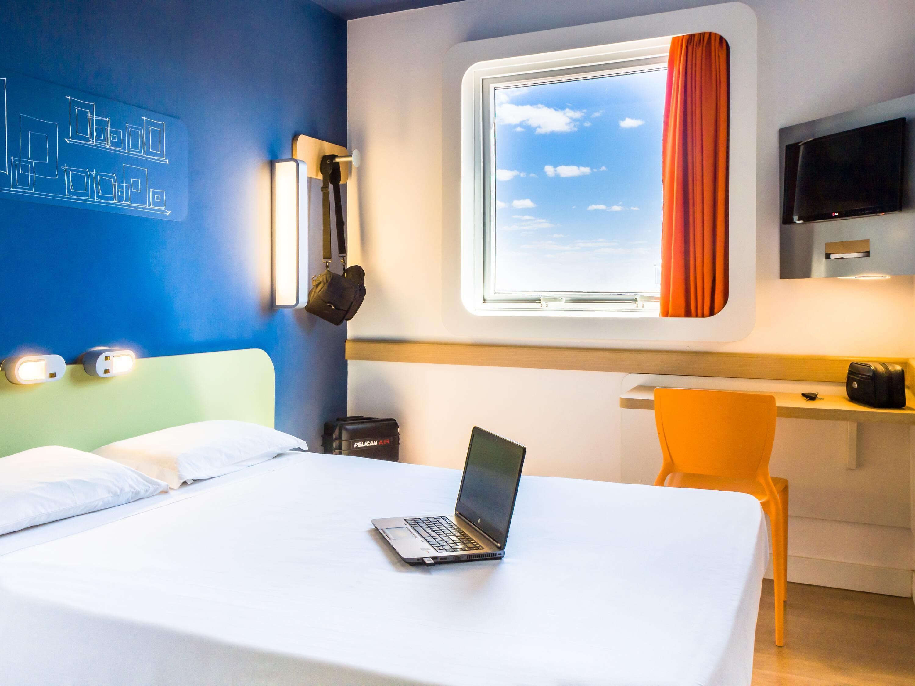 A hotel room with a bed, a desk, a laptop, a TV, and a window with curtains.