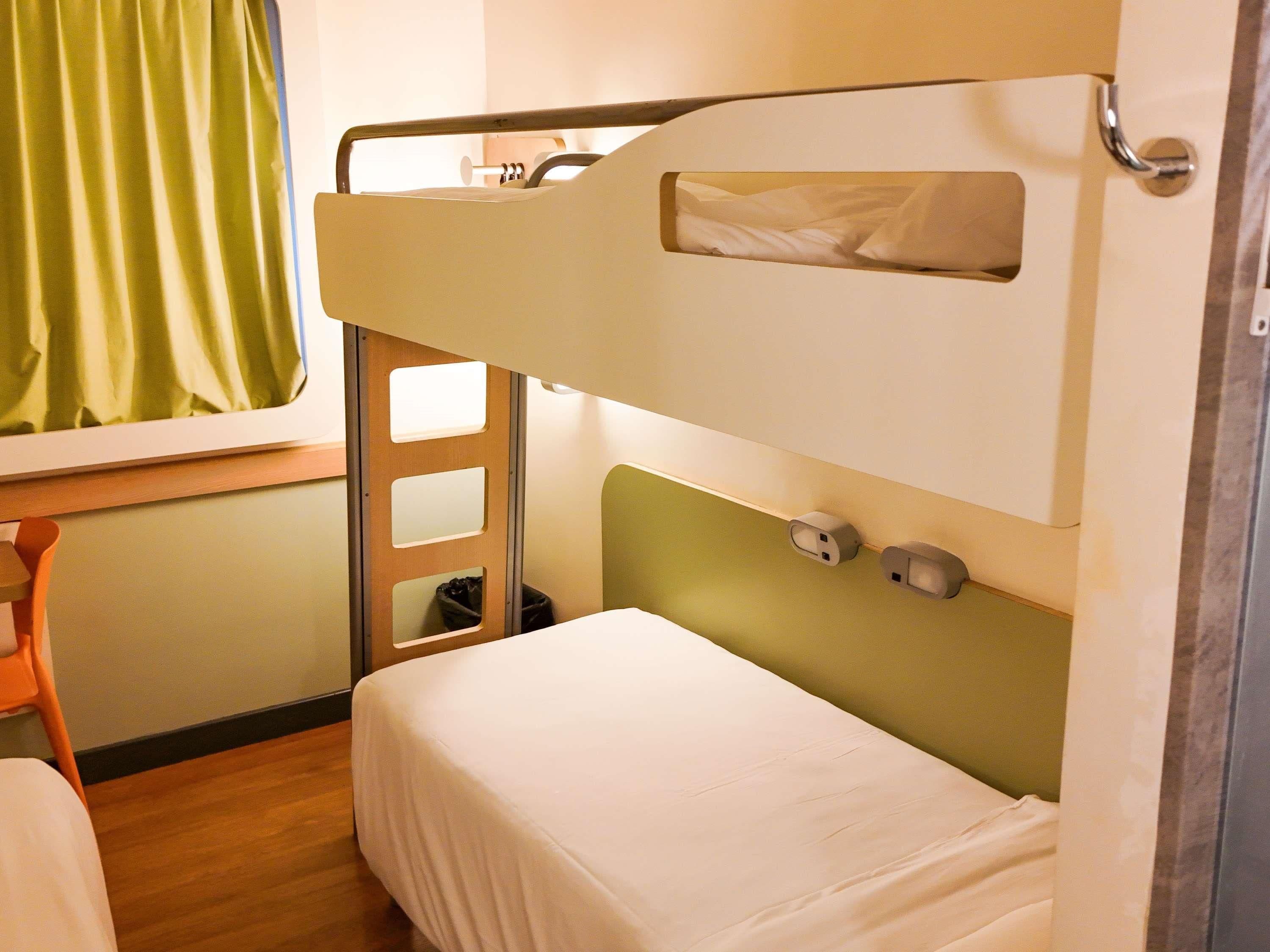 A bunk bed with a small window, a chair, and a trash bin.