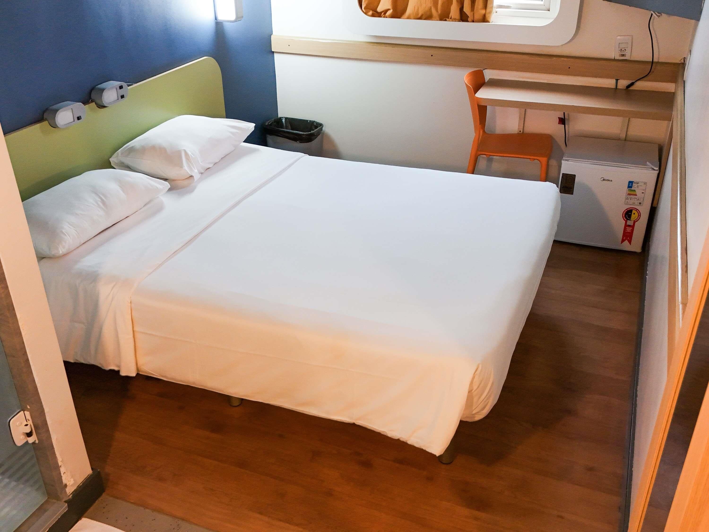 Quarto com cama, travesseiros, escrivaninha, cadeira, lixeira e geladeira.