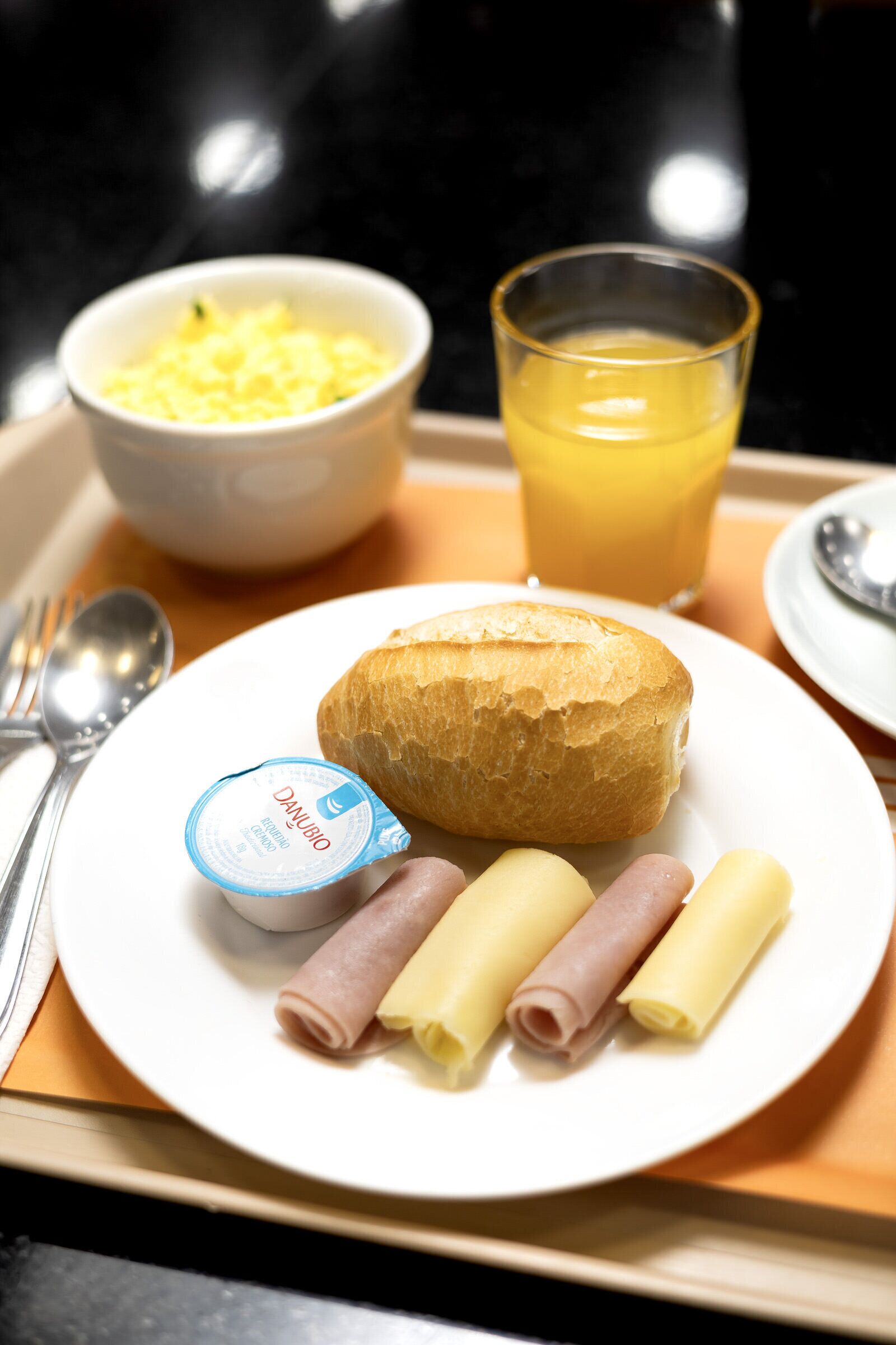 Uma bandeja de café da manhã com um croissant, mini sanduíches de presunto, um pote pequeno de iogurte, ovos mexidos e um copo de suco de laranja.
