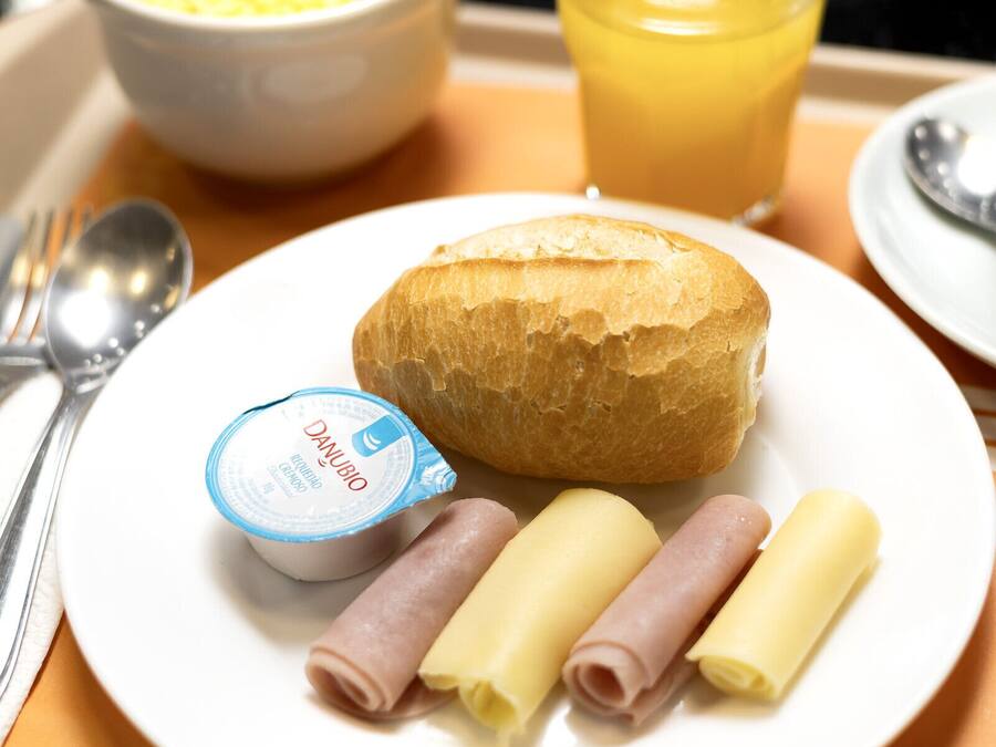 Uma bandeja de café da manhã com um croissant, mini sanduíches de presunto, um pote pequeno de iogurte, ovos mexidos e um copo de suco de laranja.