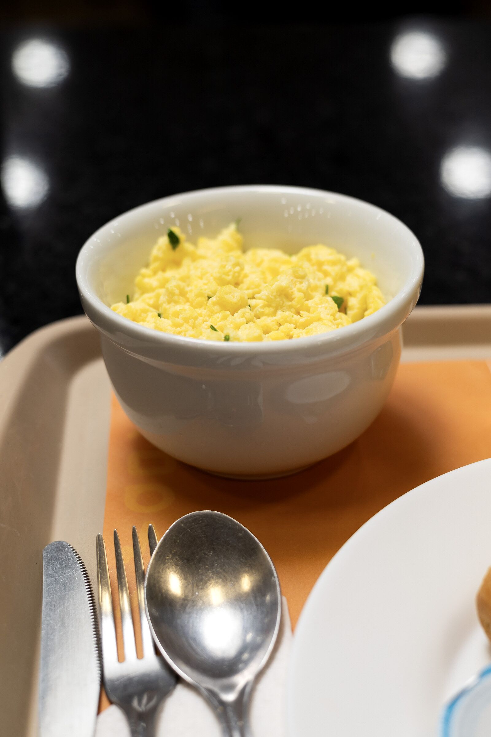 A white bowl of scrambled eggs on a beige tray with a fork and spoon beside it.