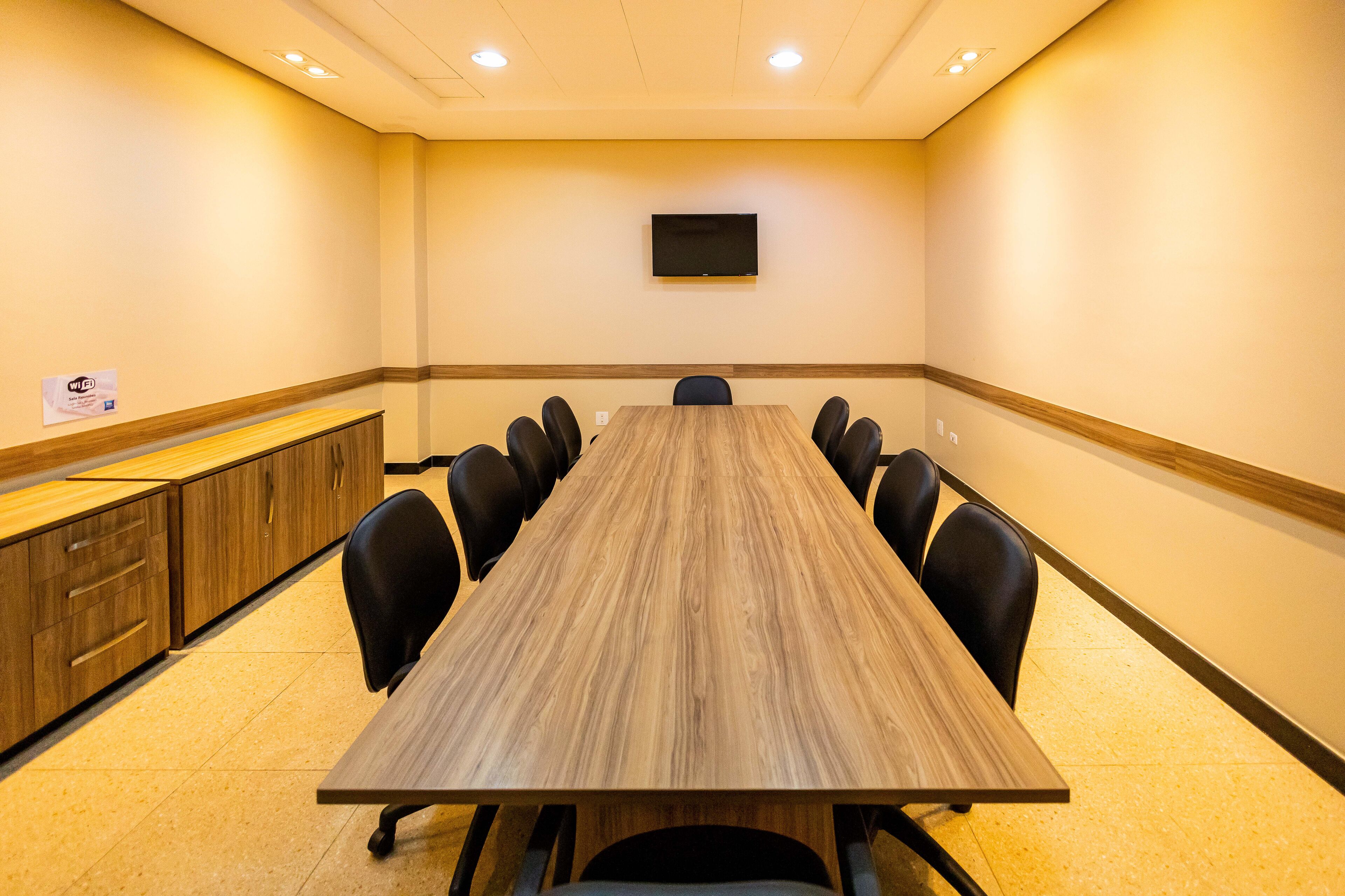 Una sala de conferencias con una larga mesa, sillas negras, un televisor en la pared y armarios de madera.