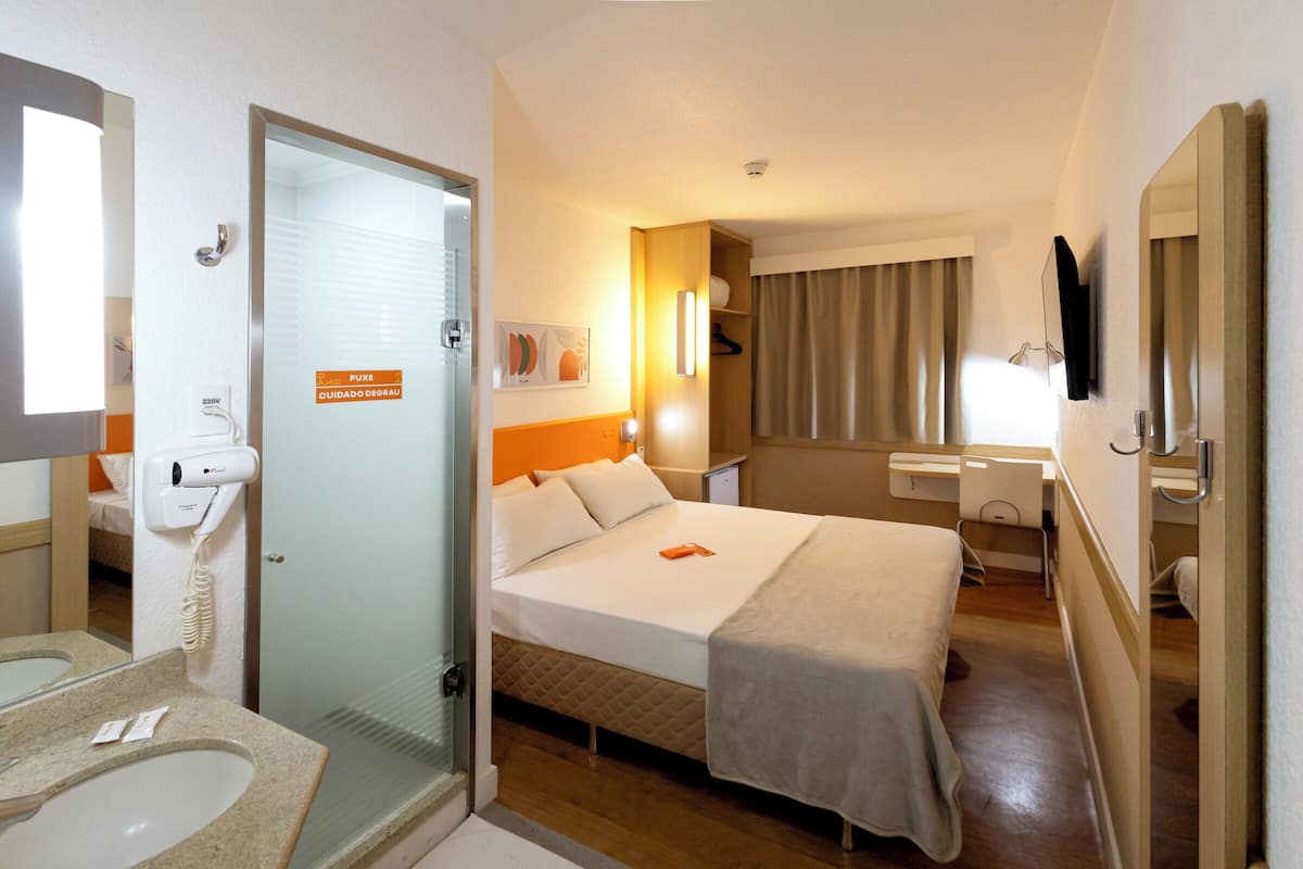 Una habitación de hotel con una cama, un baño con lavamanos y espejo, y una puerta que da a otra habitación.