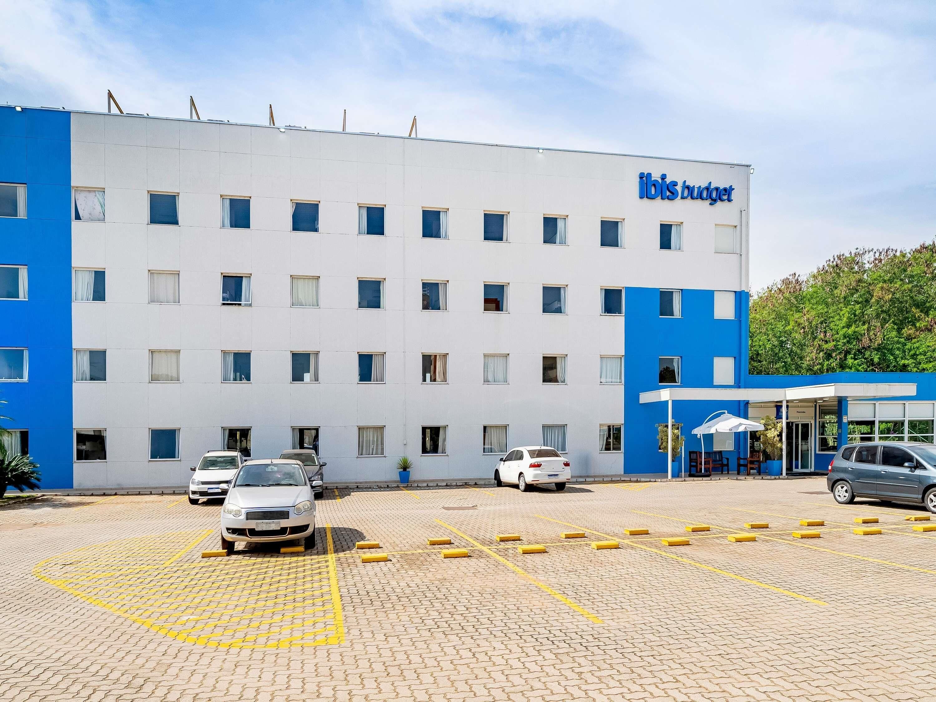 Un hotel con fachada blanca y azul, un estacionamiento con espacios delimitados y la entrada del edificio con un toldo.