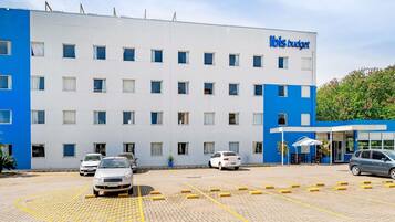Un hotel con fachada blanca y azul, un estacionamiento con espacios delimitados y la entrada del edificio con un toldo.