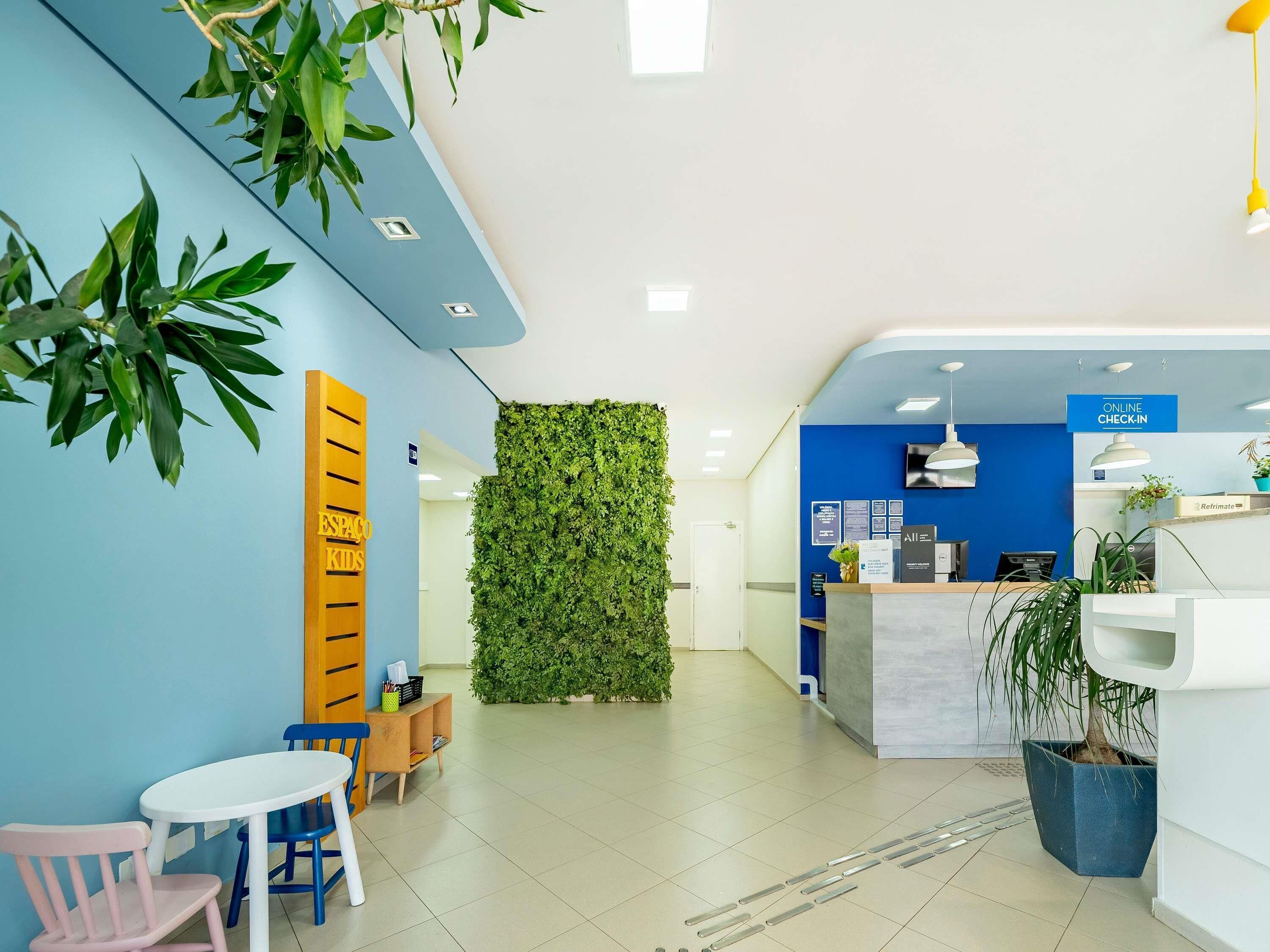 Een receptie met een blauwe wand, een groene verticale tuin, een receptiebalie en een kinderspeelruimte met een roze stoel en een witte tafel.
