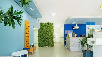 Een receptie met een blauwe wand, een groene verticale tuin, een receptiebalie en een kinderspeelruimte met een roze stoel en een witte tafel.