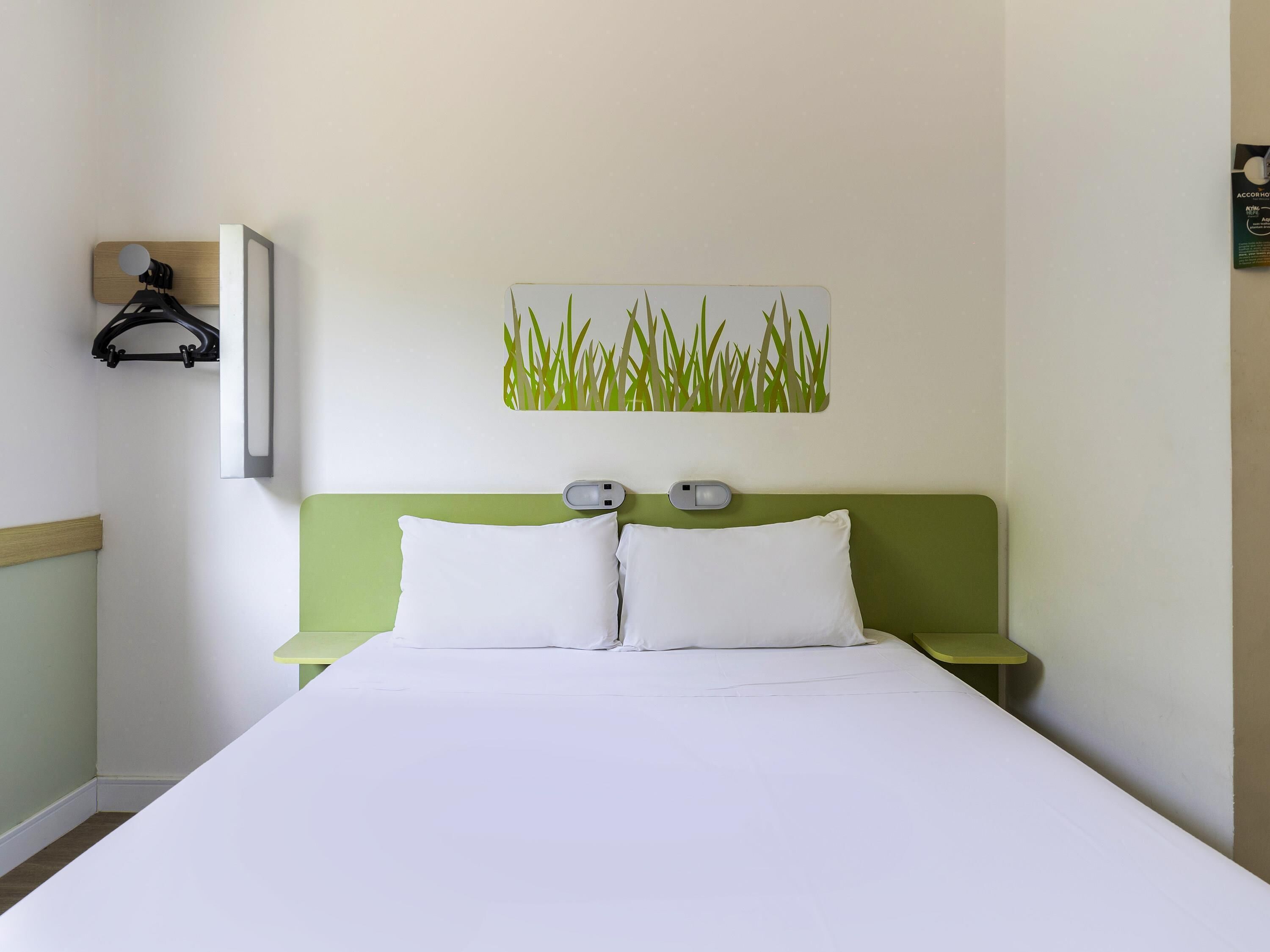 Habitación de hotel con cama, cabecera verde, almohadas blancas, lámpara fijada a la pared y una obra de arte con motivos de hierba.