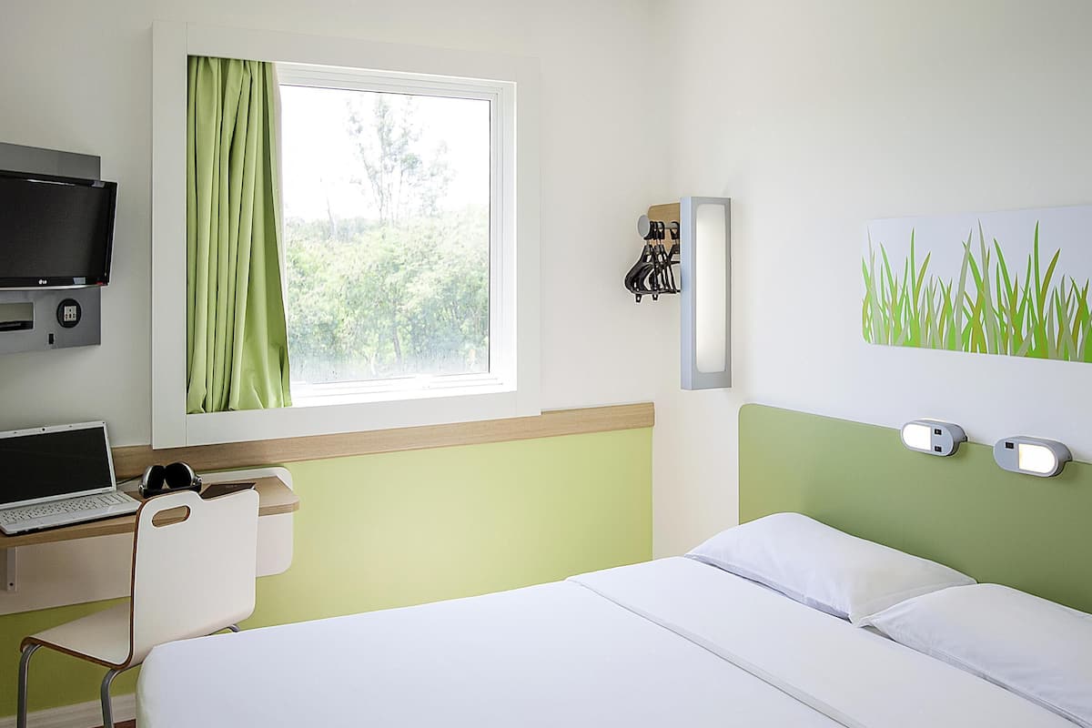 Una habitación de hotel con una cama, un escritorio con una computadora portátil, un televisor y una ventana con cortinas.