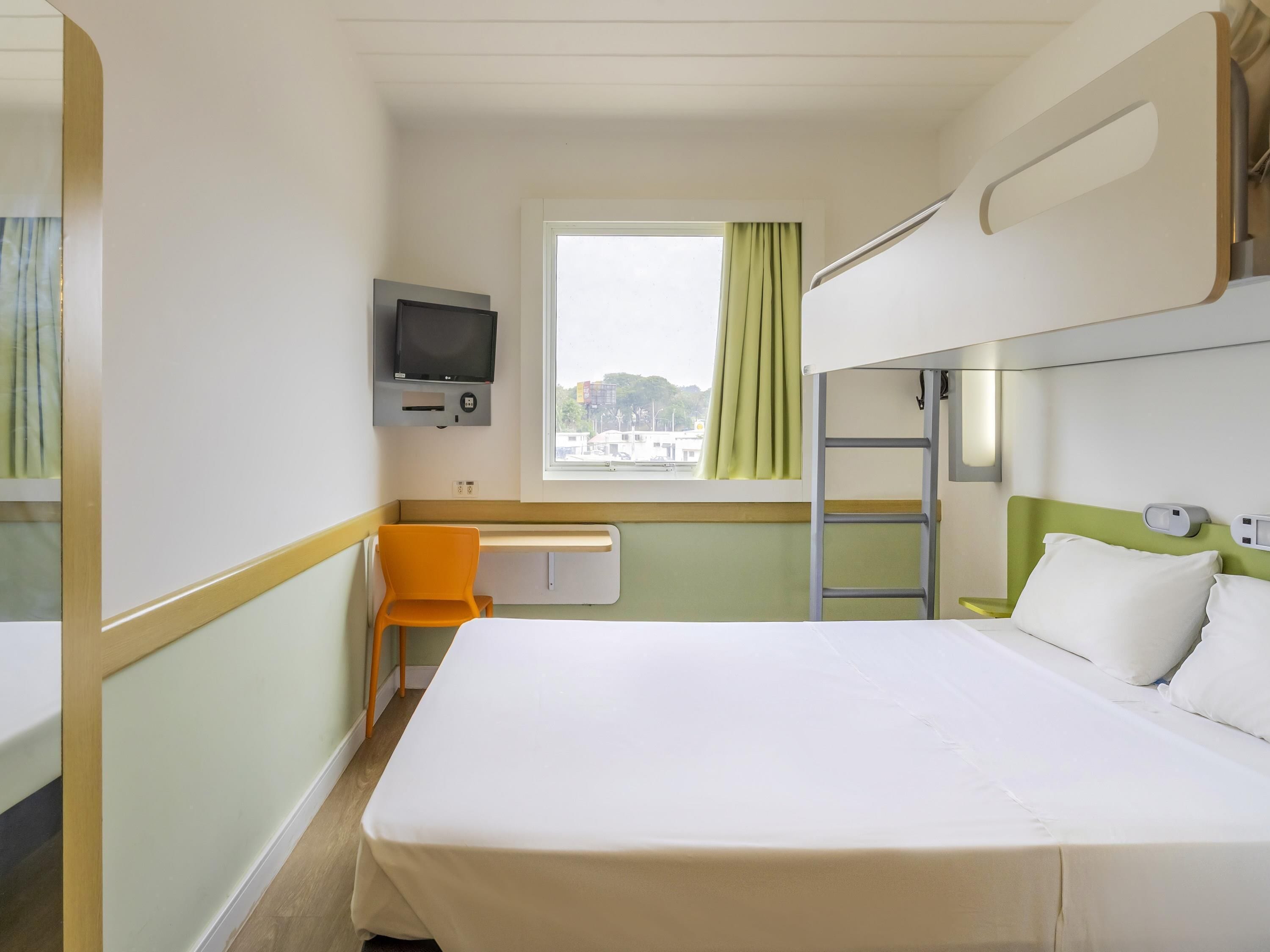 Une chambre d’hôtel avec un lit superposé, un bureau avec une chaise, une télévision, une fenêtre avec des rideaux et une vue sur la ville.