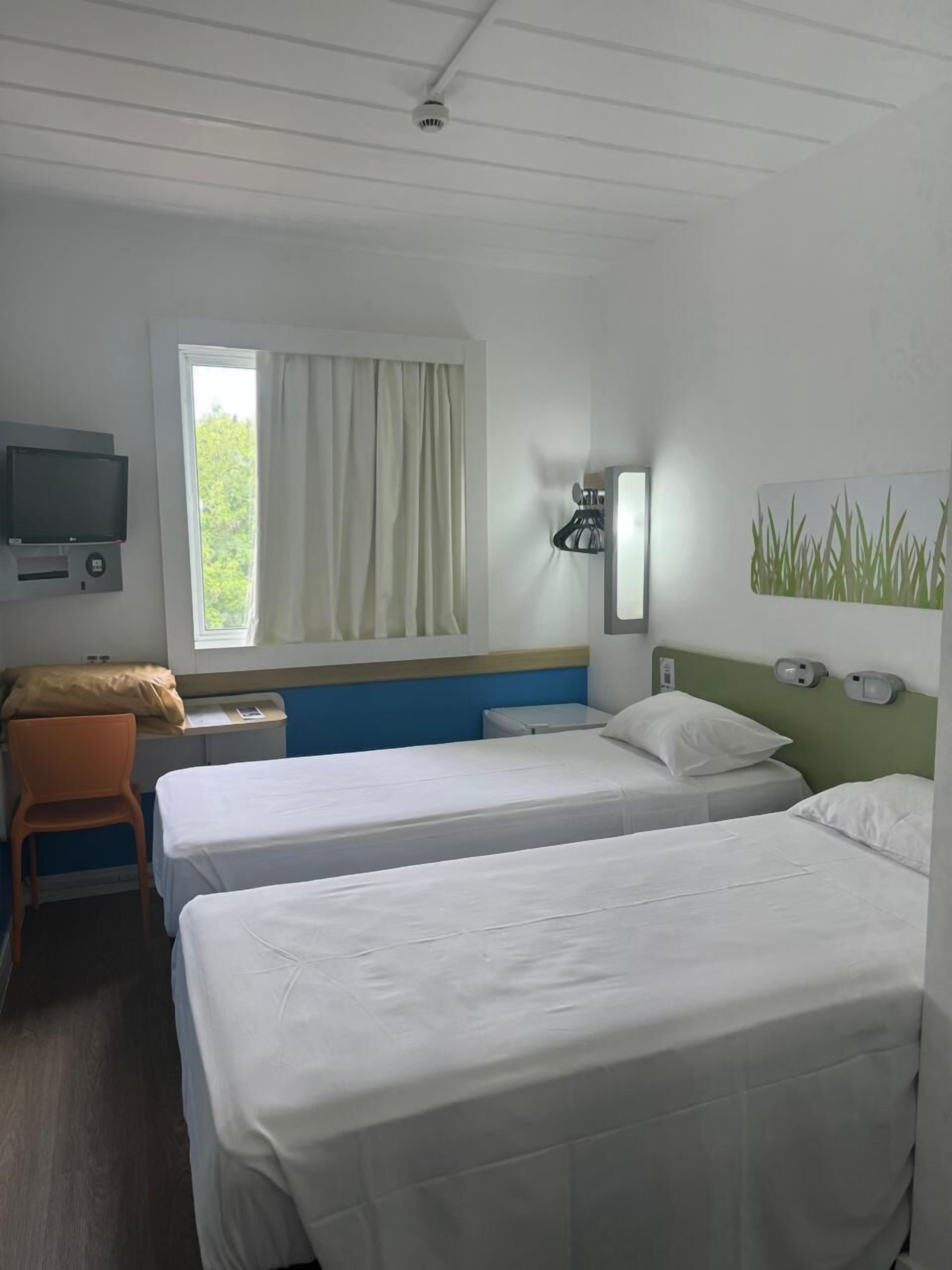Quarto de hotel com duas camas, uma mesa com cadeira, televisão, janela com cortinas e luminária na parede.