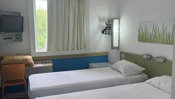 Quarto de hotel com duas camas, uma mesa com cadeira, televisão, janela com cortinas e luminária na parede.