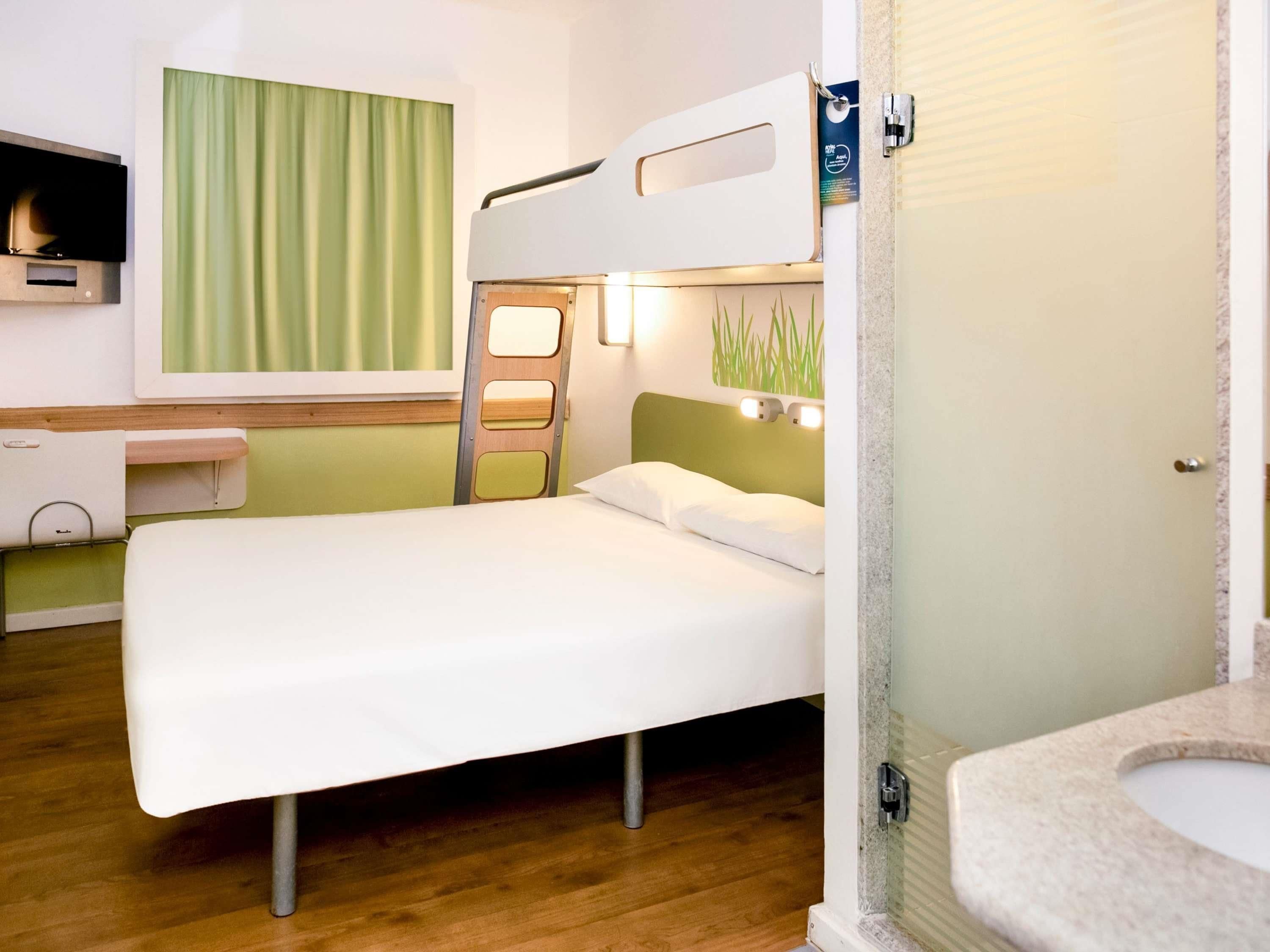 A single-bed room with a bunk bed, a desk, a television, a bathroom with a sink, and a green curtain.