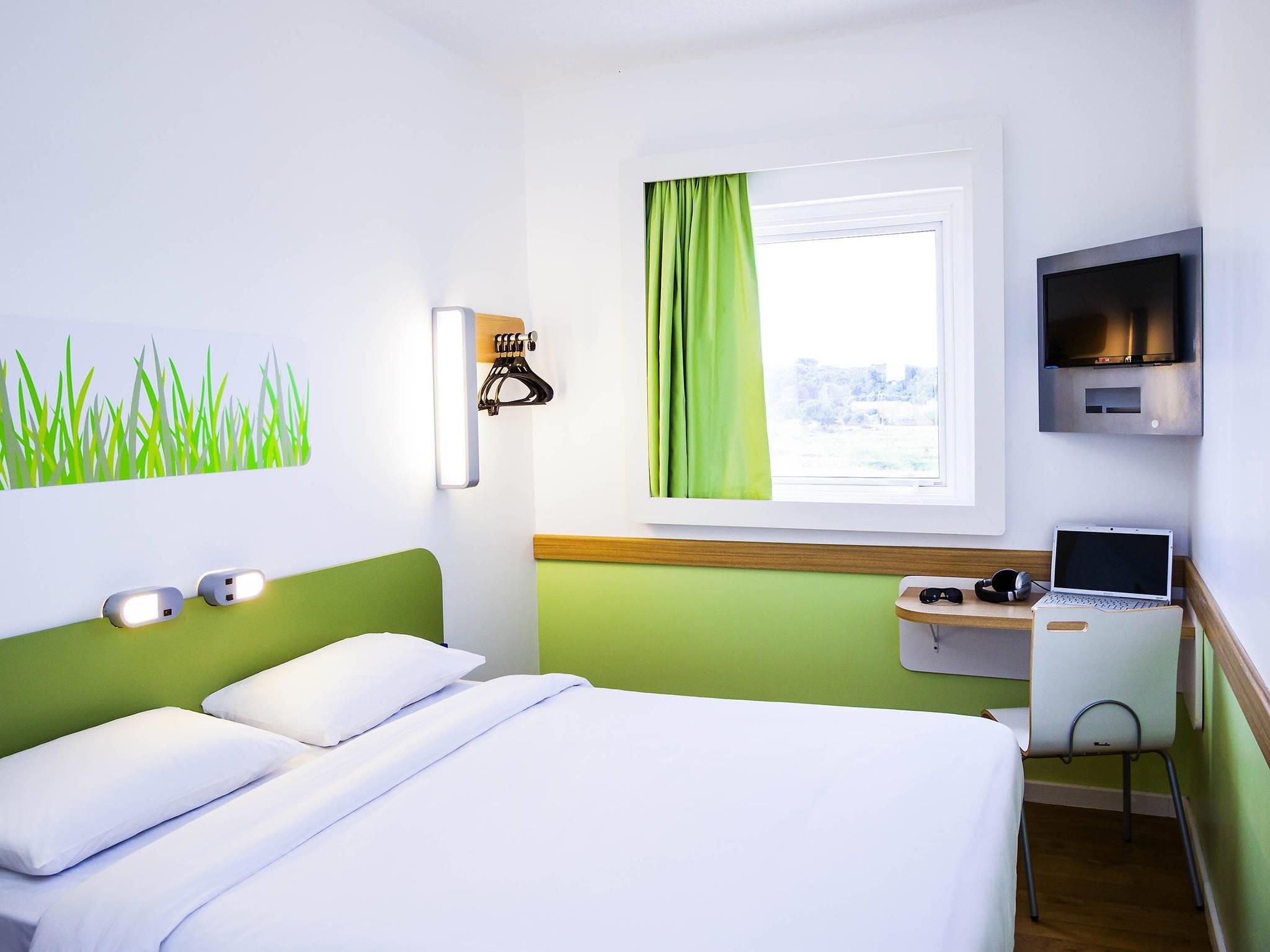 A hotel room with a bed, a desk with a laptop, a TV, and a window with curtains.