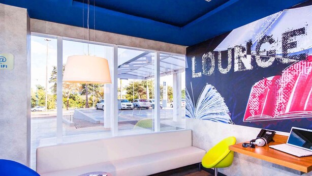 Un'area lounge moderna con un'ampia finestra, un divano, un tavolo di legno con un portatile e un murales colorato al muro.