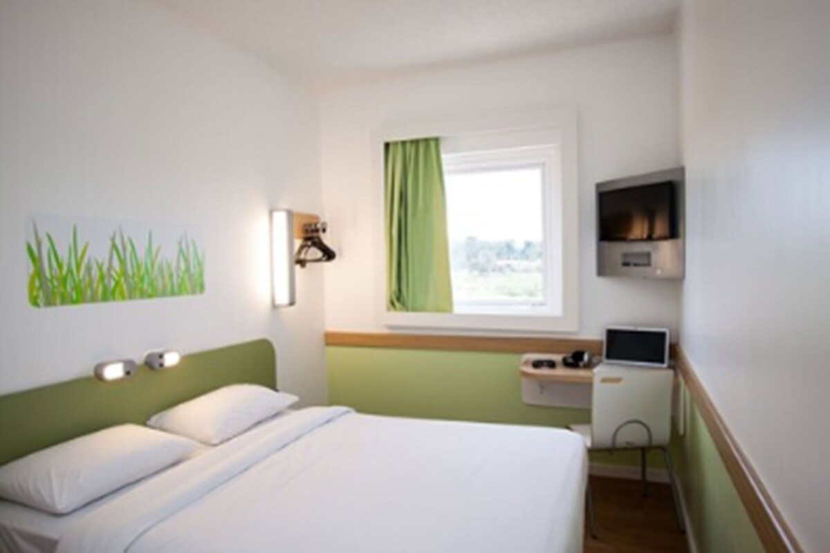 A hotel room with a bed, a desk with a computer, a TV, and a window with a view.