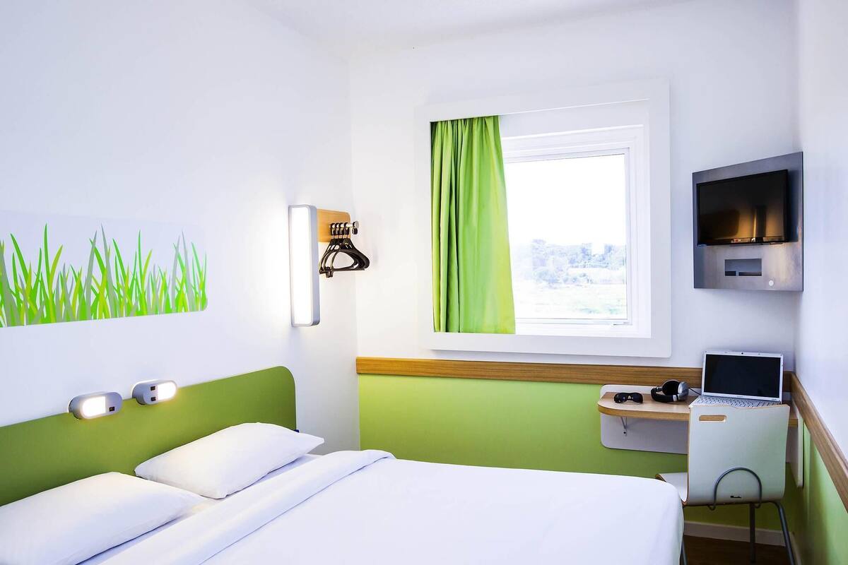 Une chambre d’hôtel avec un lit, un bureau avec un ordinateur portable, une télévision et une fenêtre avec des rideaux.
