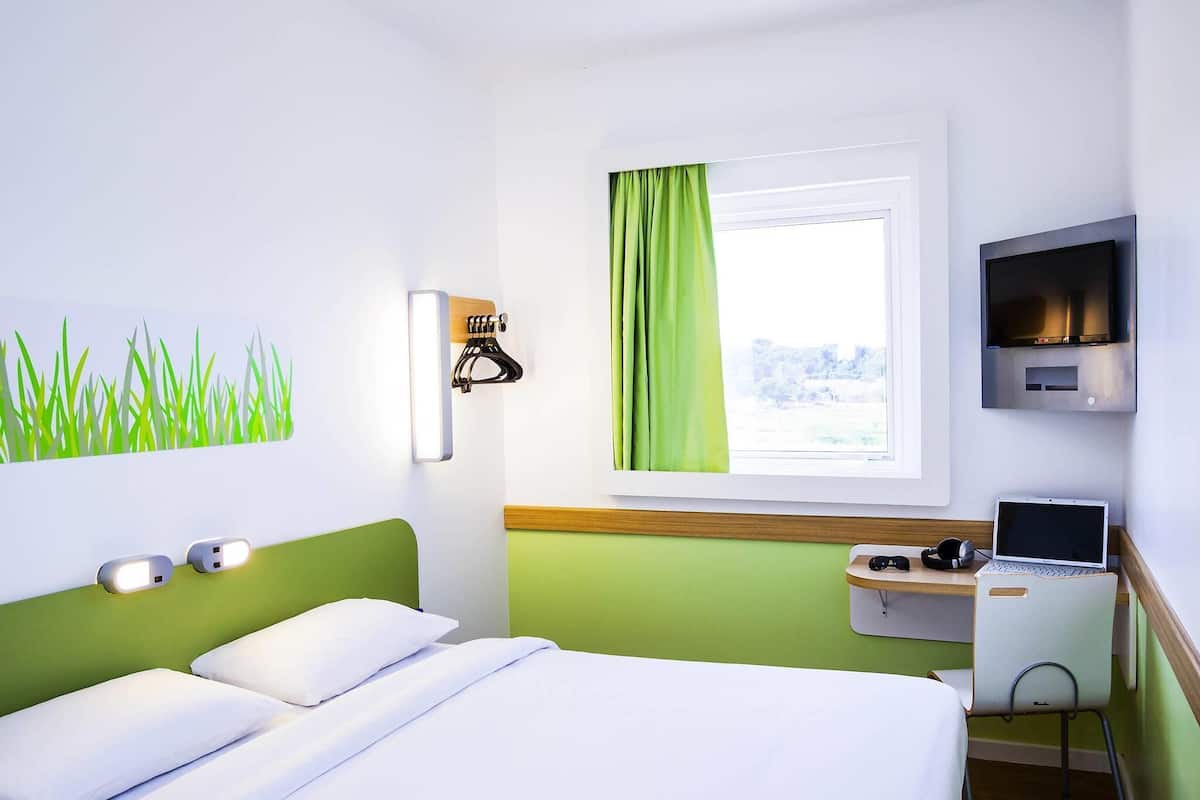 A hotel room with a bed, a desk with a laptop, a TV, and a window with curtains.