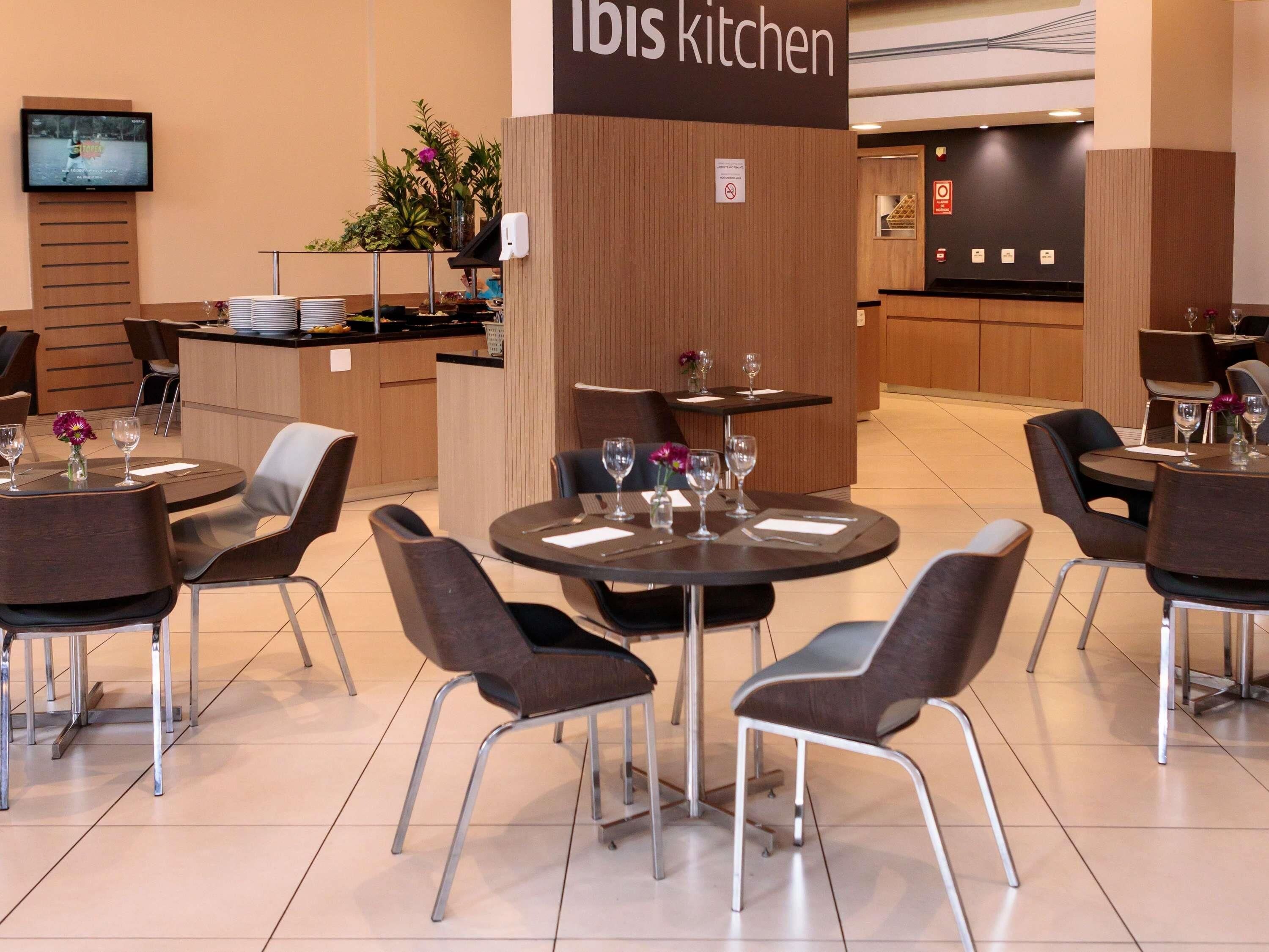 Interior moderno de restaurante com mesas redondas, cadeiras e uma área de cozinha identificada como 'Ibis kitchen'.