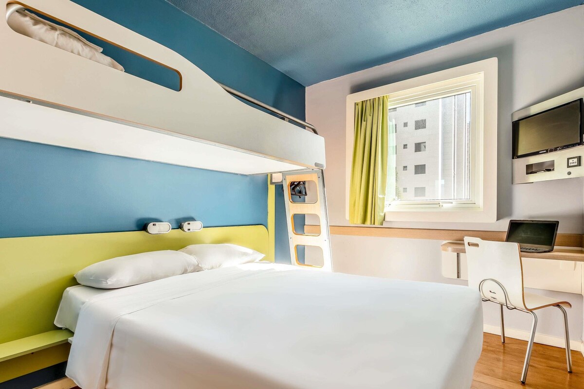 A hotel room with a bunk bed, a desk with a laptop, a TV, and a window with curtains.