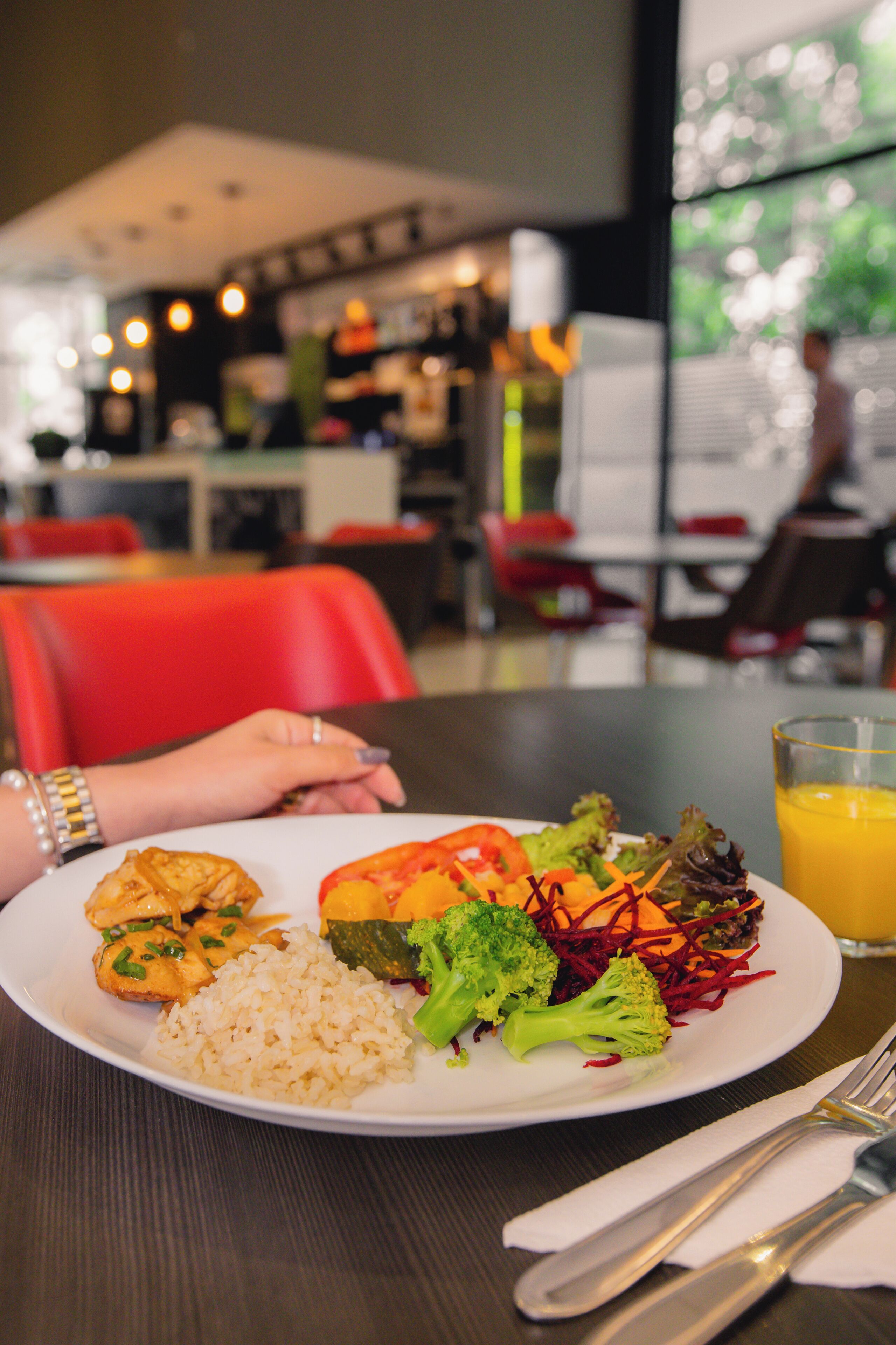 Une assiette de nourriture contenant du riz, des légumes et du poulet, un verre de jus d’orange et la main d’une personne portant une bague, le tout sur une table dans un restaurant.