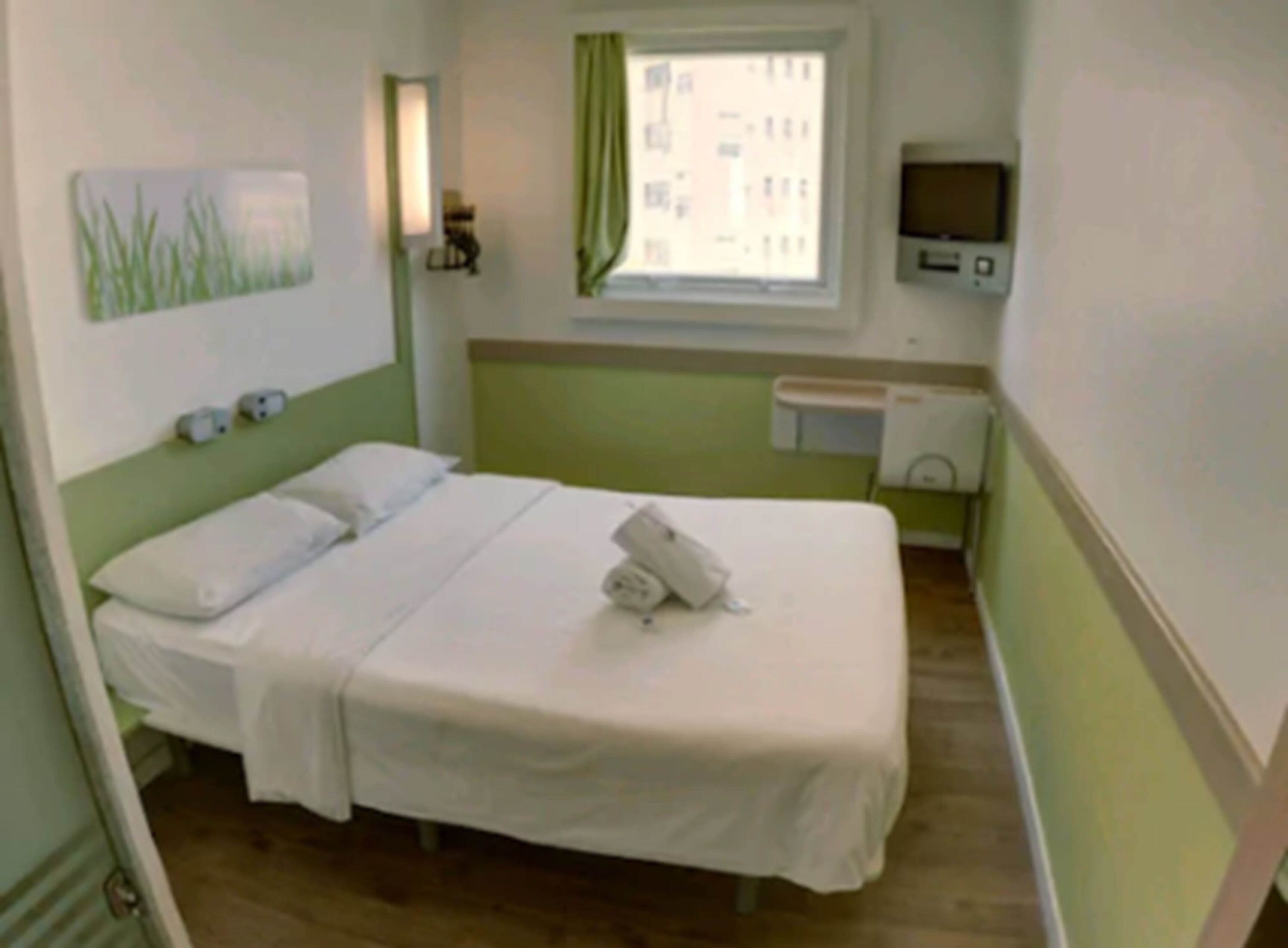 Una habitación pequeña y limpia con cama, ventana con cortinas verdes, televisor en la pared y una mesita con calefactor.