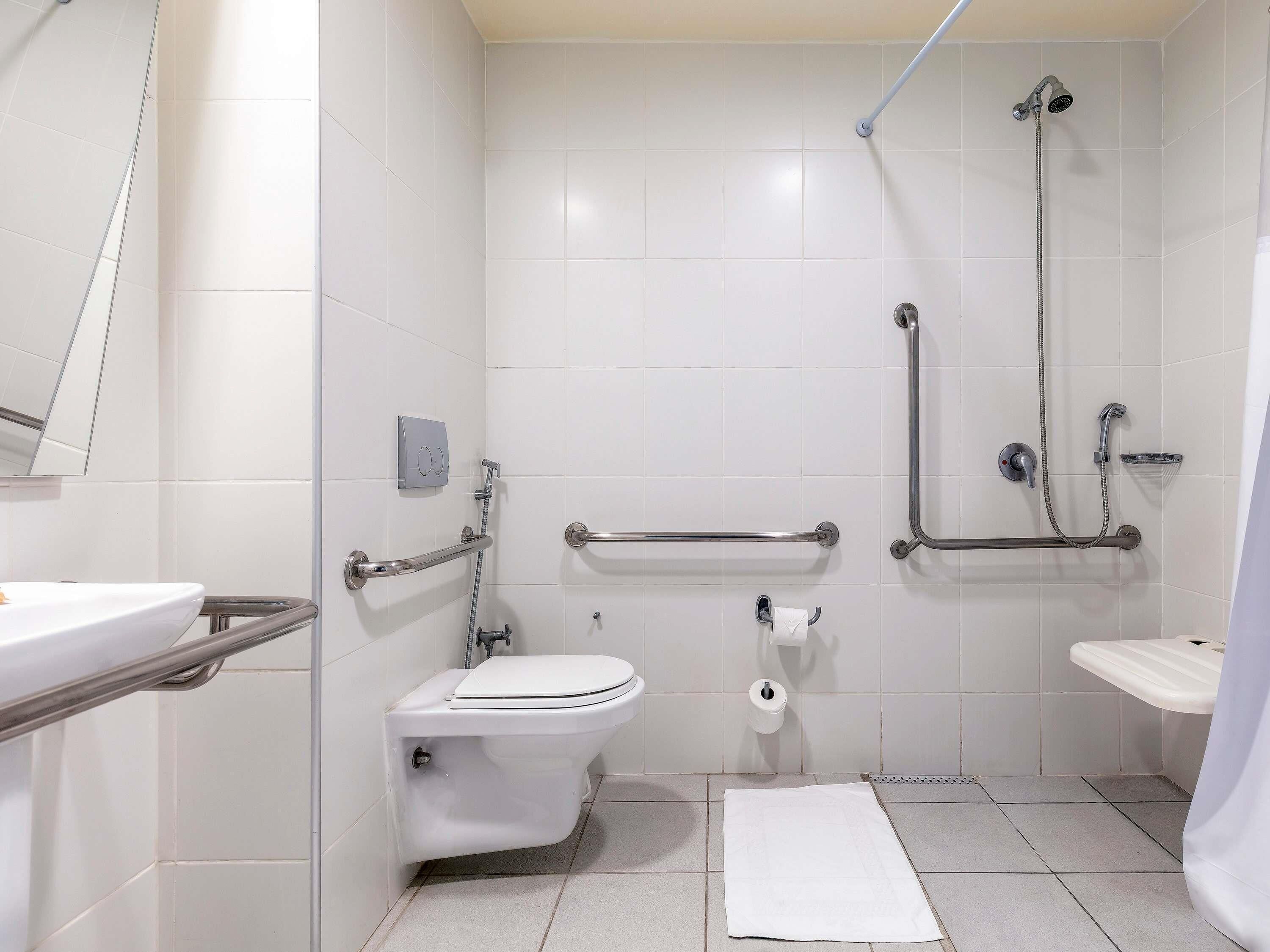 Une salle de bain carrelée de blanc, avec des toilettes, un lavabo, une douche équipée de barres d’appui et une serviette sur le sol.