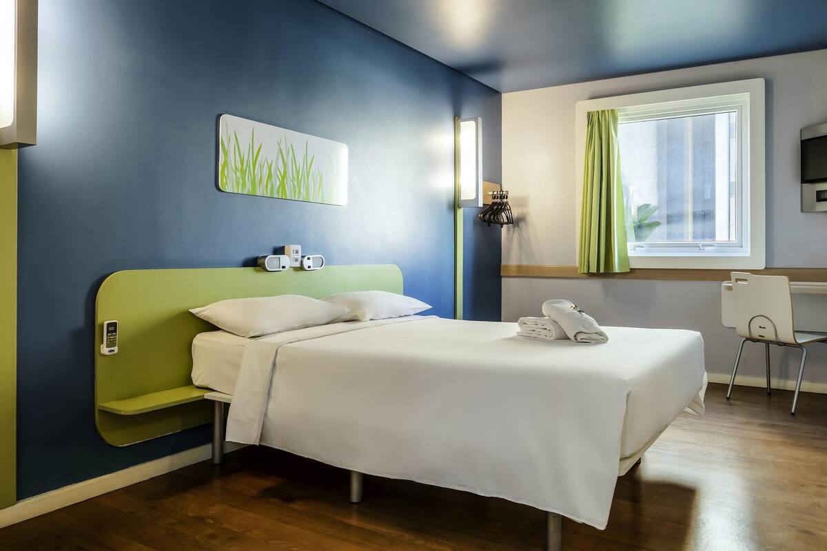 A hotel room with a bed, bedside tables, a desk, a chair, a window with curtains, a wall-mounted TV, and a painting.