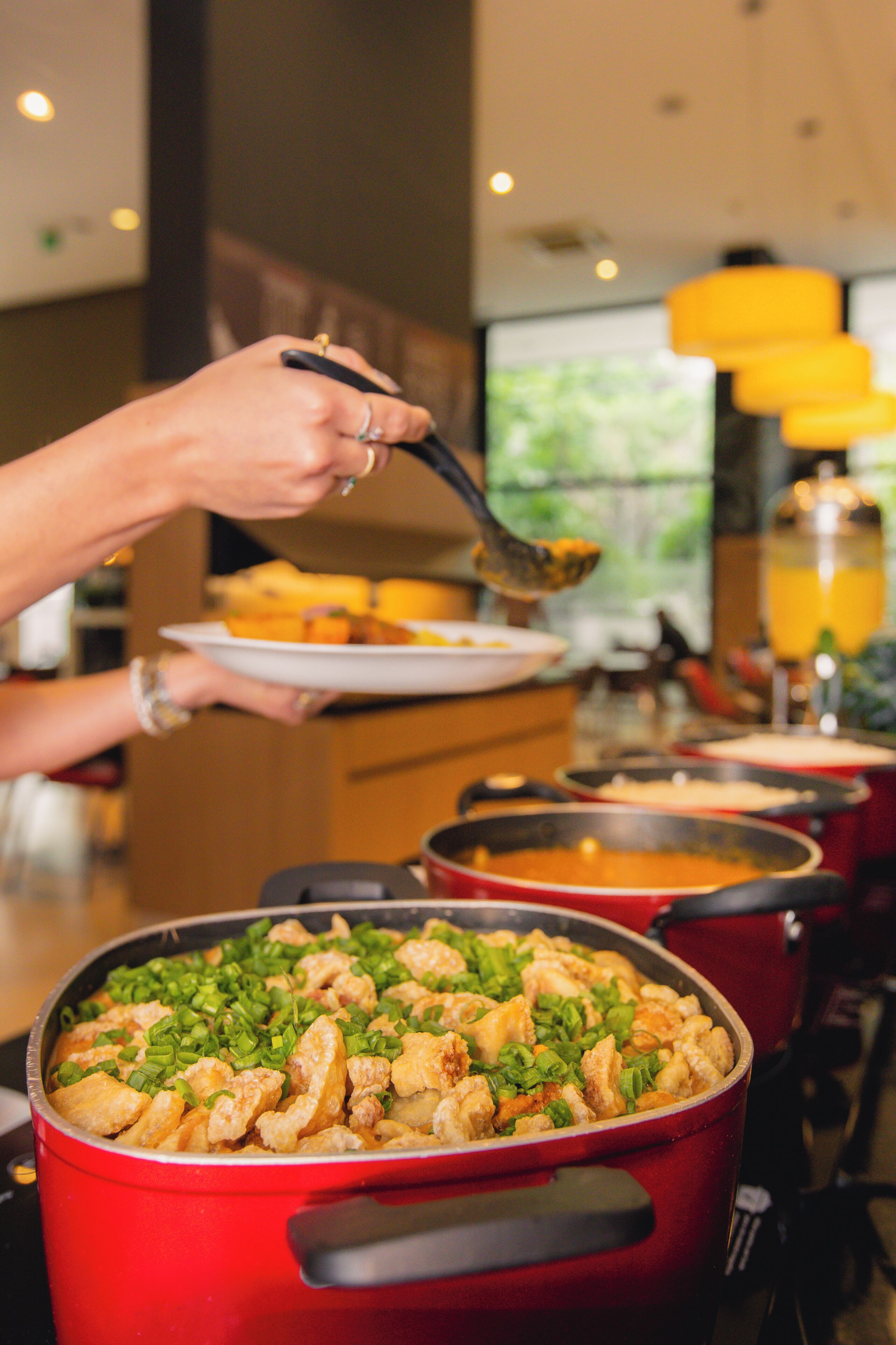 A buffet with various dishes, including a red pot with a green garnish, and a person serving food with a spoon.